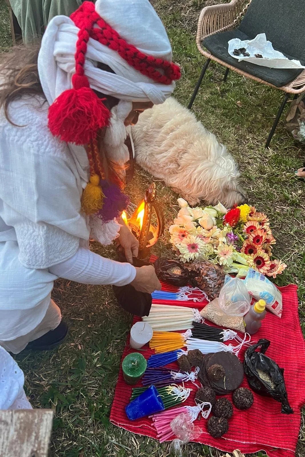 Person wearing a white outfit and a red and white cloth head covering with colorful tassels, holding a small torch, participating in a fire ritual outdoors. Next to them is a white dog lying on the ground. In front of them, there is a red cloth with flowers, candles, incense sticks, and various ritual items on it.