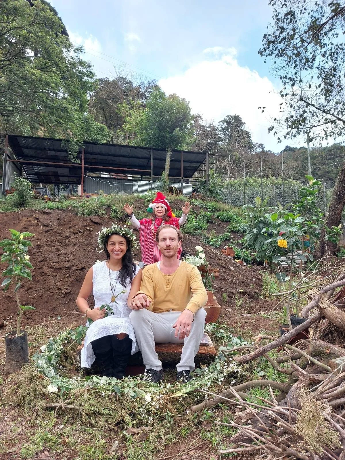 Three people sitting on the ground in a garden, with a woman standing behind them. The woman is dressed as Santa Claus, and the woman sitting in front has a floral crown. The man is in a yellow shirt. The area is decorated with white flowers and rustic elements, and the background shows trees, plants, and a structure with a roof.