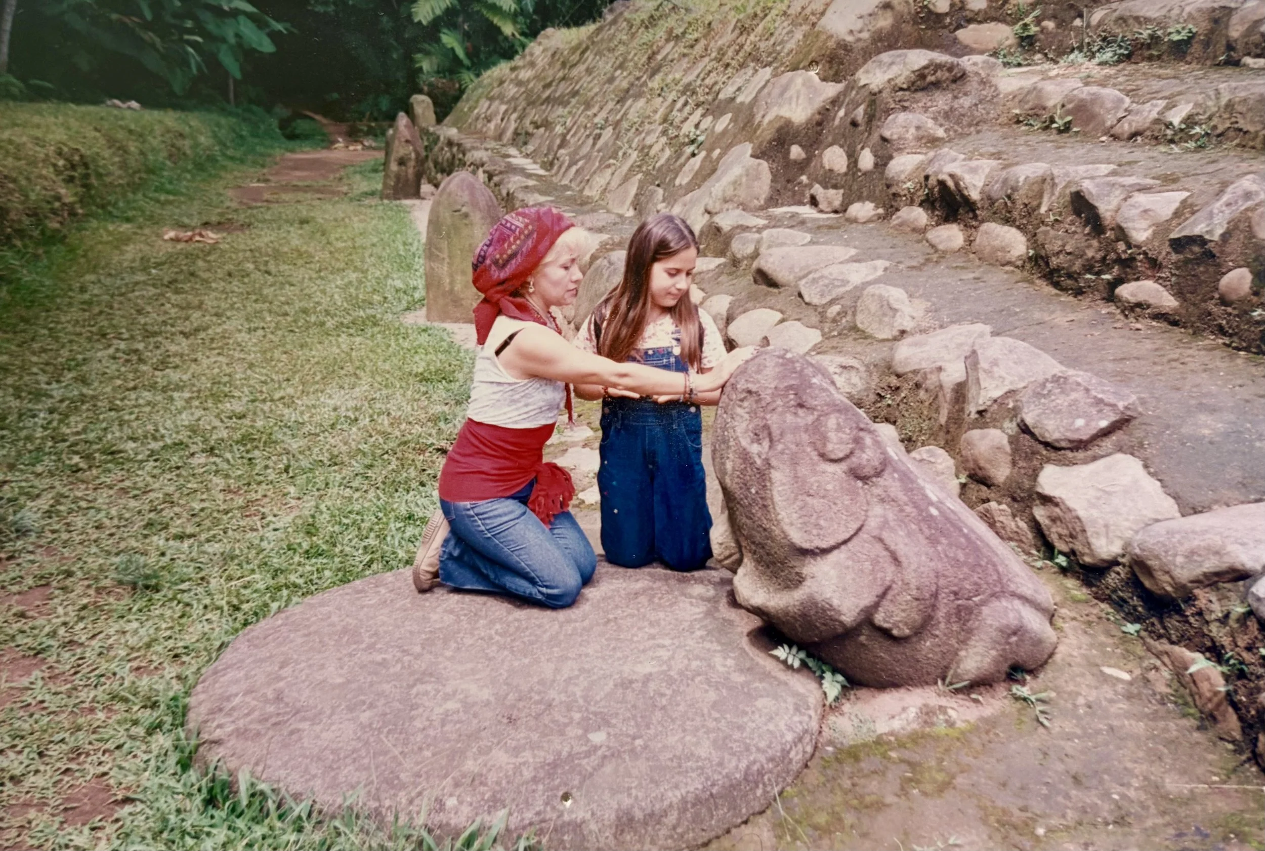 A woman and a young girl kneeling on the grass in front of an ancient stone sculpture, touching it, with stone steps and greenery in the background.