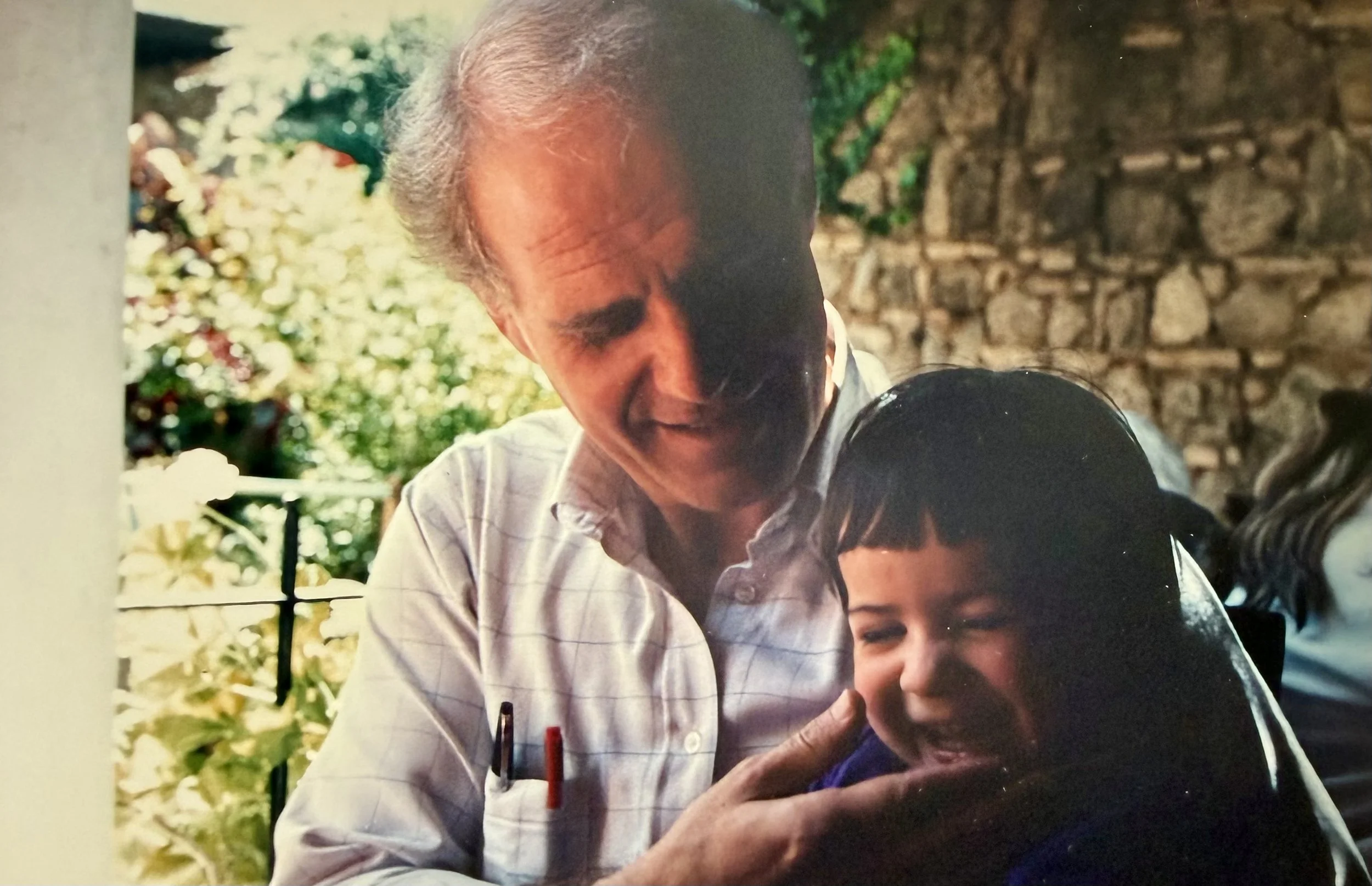 An older man with gray hair smiling and holding a young girl with dark hair and bangs, who is also smiling and looking down.