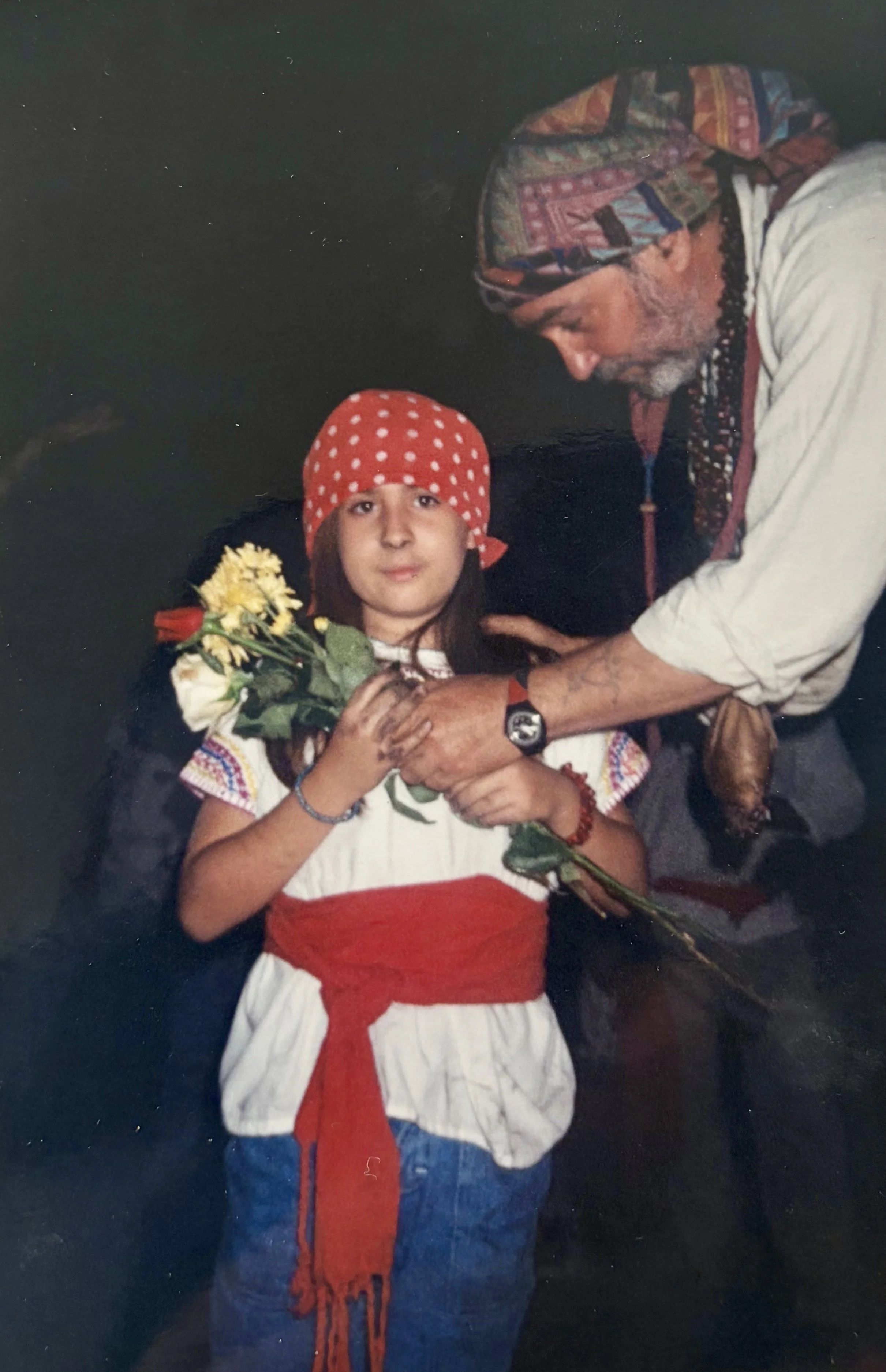 A young girl with brown hair, wearing a red and white polka dot headscarf, holding flowers, beaded jewelry, and a flower, standing next to an older man with a gray beard, wearing a colorful headscarf and traditional clothing, in a dimly lit setting.