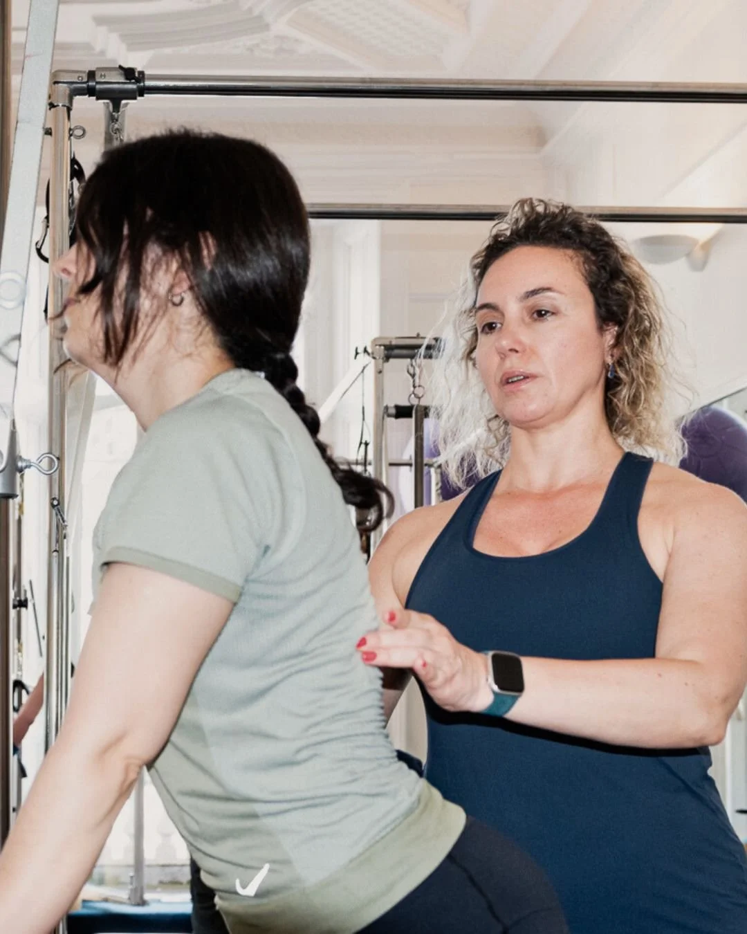 A woman in a dark blue tank top assisting another woman with a fitness or rehabilitation exercise using a Pilates or fitness reformer machine.