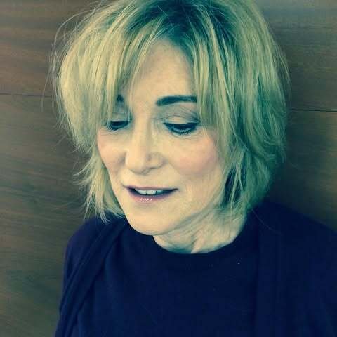 Woman with short blonde hair and wearing a dark shirt, looking down, indoors with wooden panel background.