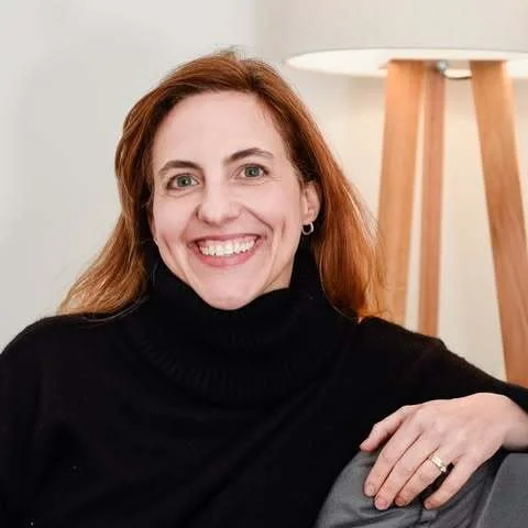 A woman with long red hair smiling, wearing a black turtleneck sweater, sitting indoors next to a wooden floor lamp.
