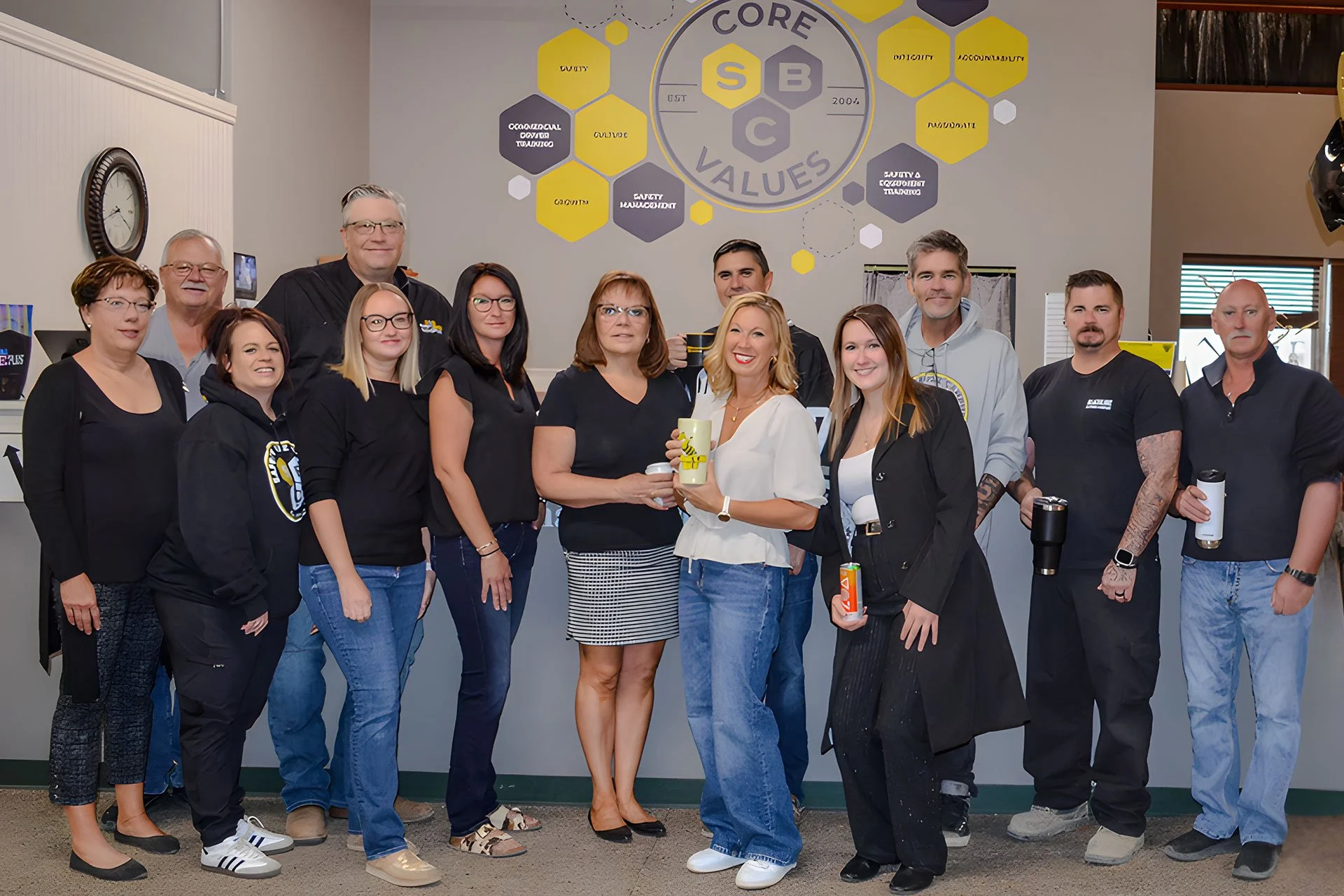A group of fourteen people standing inside a room with a core values chart on the wall behind them. The group includes men and women of various ages, some holding cups or water bottles, and smiling at the camera.