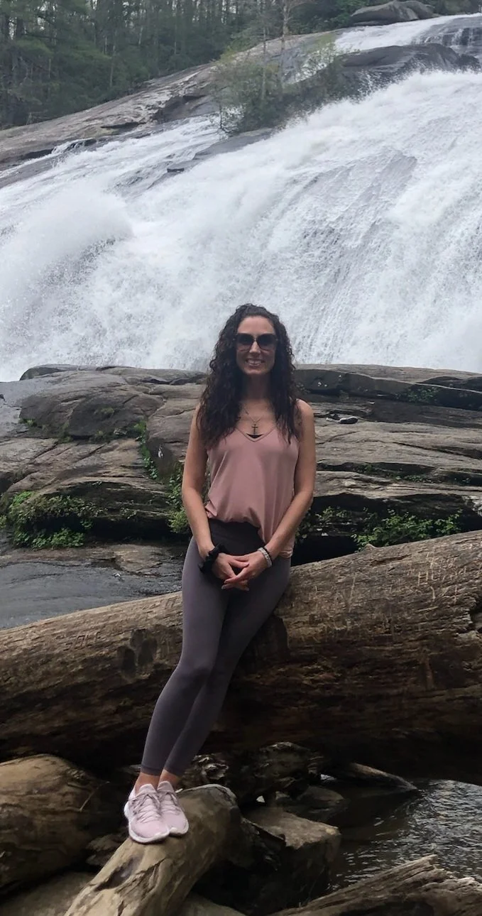 A woman with curly dark hair wearing sunglasses, a pink sleeveless top, gray leggings, and pink sneakers, standing on a large fallen log in front of a waterfall in a forested area.