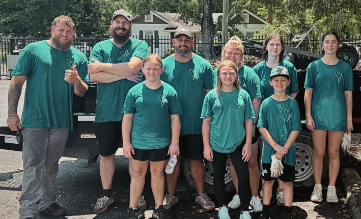 Group of nine people, including children and adults, wearing matching teal shirts with a heart logo, standing outdoors with a black fence and houses in the background.