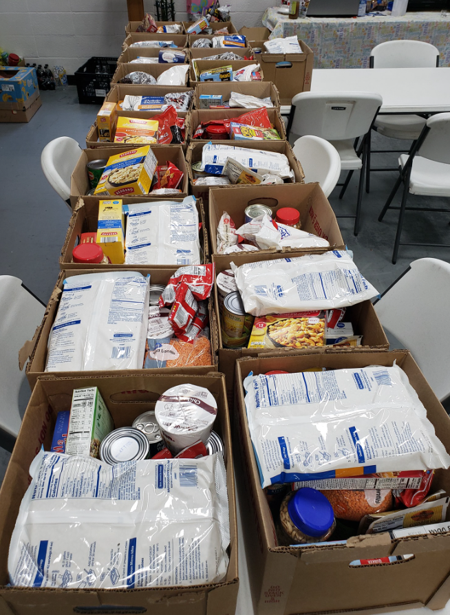 Tables filled with cardboard boxes containing packaged food items such as canned goods, dry pasta, and rice in a community center or food bank setting.