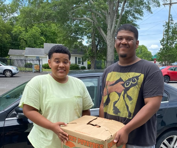 Two young men standing outdoors, holding a large cardboard box with potatoes, in front of a black car. One is wearing a light green T-shirt and the other is wearing a dark T-shirt with a graphic print.