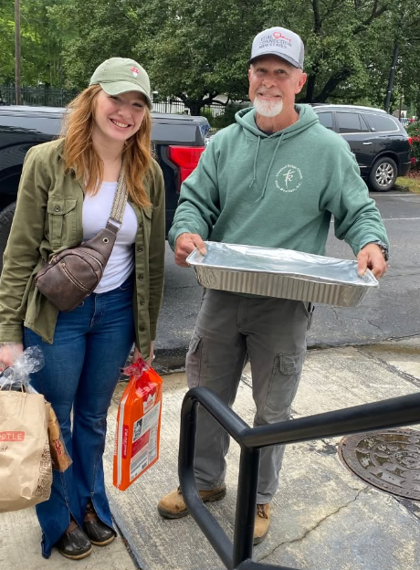 A woman and a man standing outdoors near parked cars. The woman is wearing a light green cap, green jacket, and jeans, holding a paper bag and a boxed item. The man, wearing a white cap with red writing, green hoodie, and gray pants, is smiling and holding a large aluminum tray covered with foil.