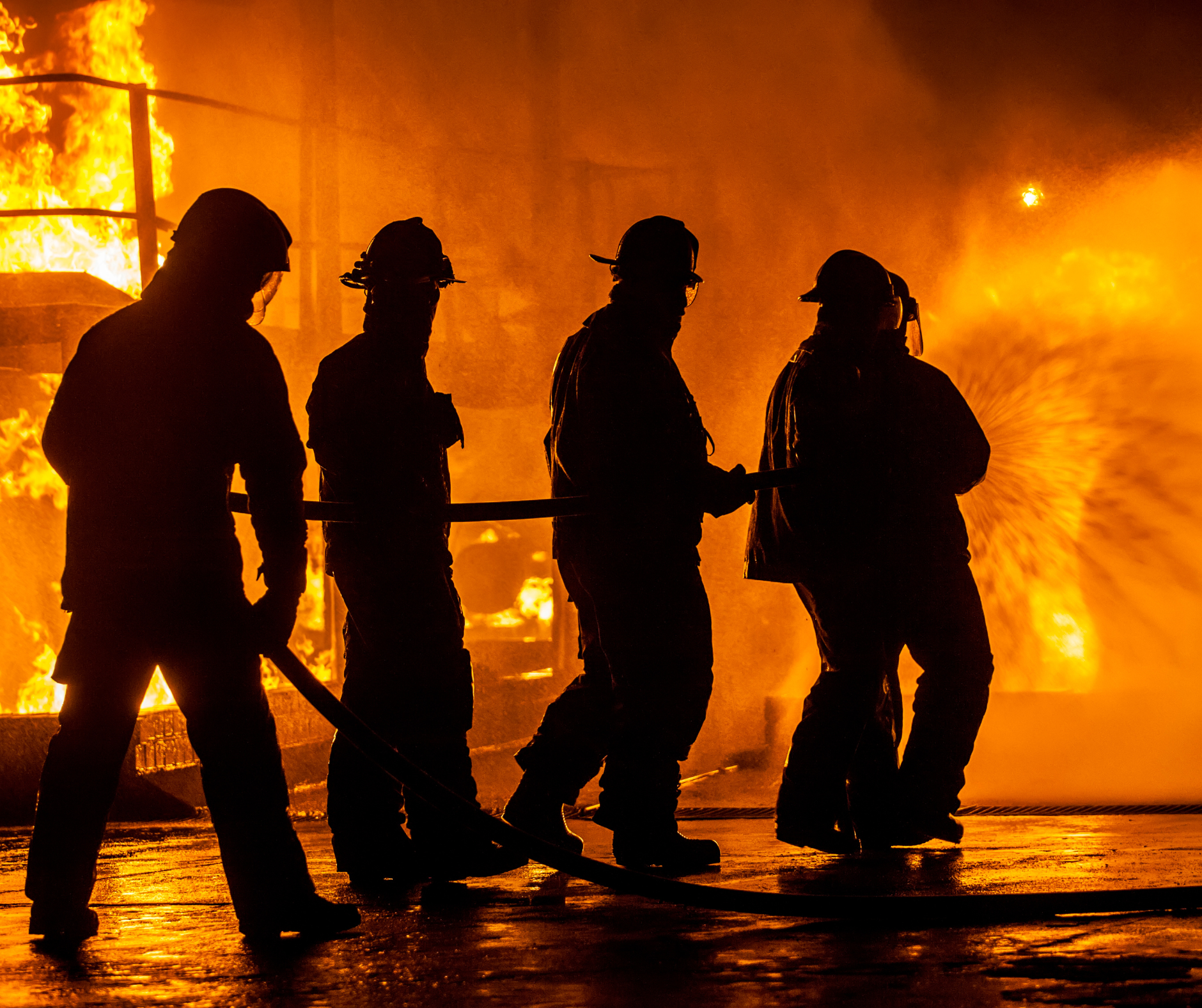 Silhouettes of four firefighters fighting a large blaze at night, with flames and smoke in the background.
