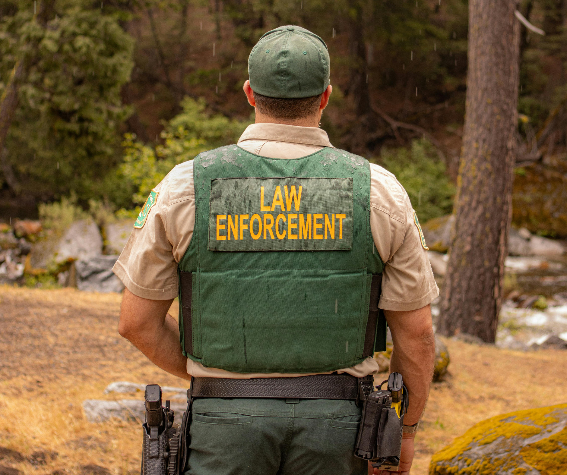 Law enforcement officer in uniform with a green vest labeled 'LAW ENFORCEMENT' facing a forested outdoor area.
