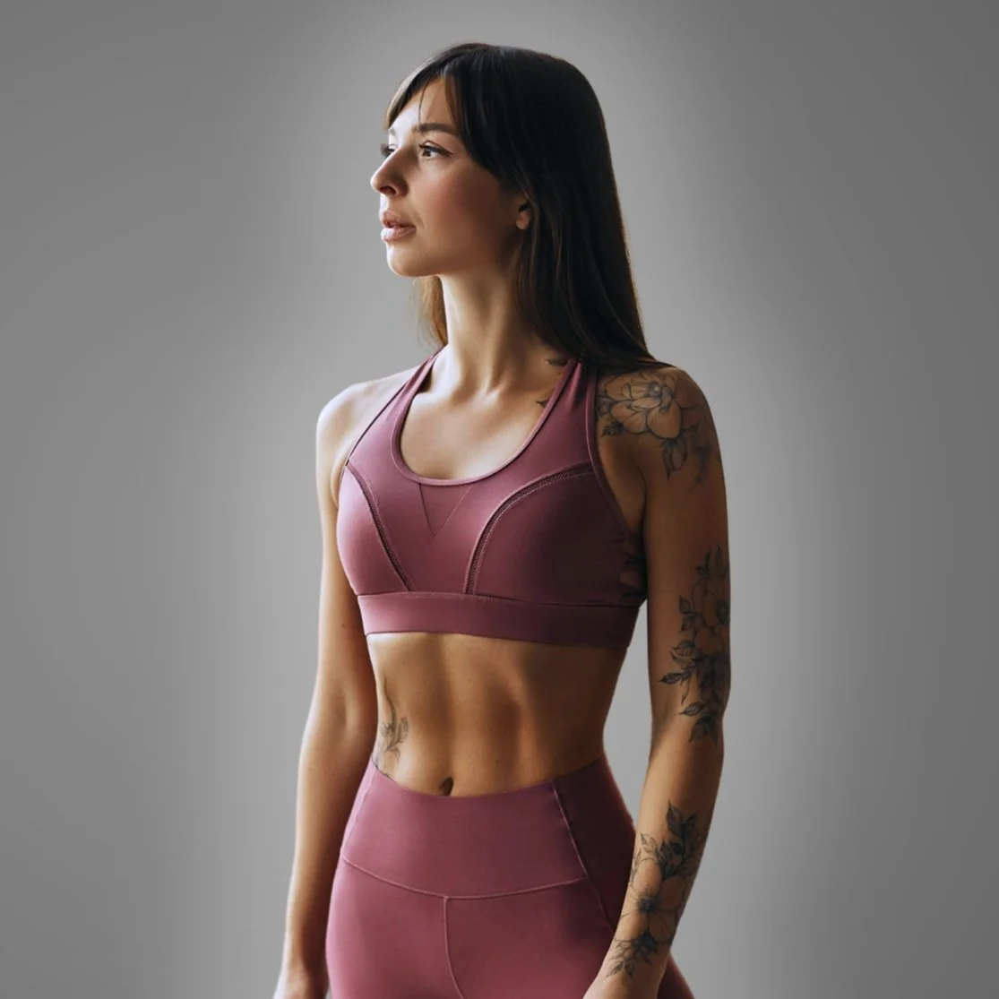 Fitness woman wearing mauve sports bra and leggings, standing sideways with tattoos on her right arm and torso, neutral gray background.