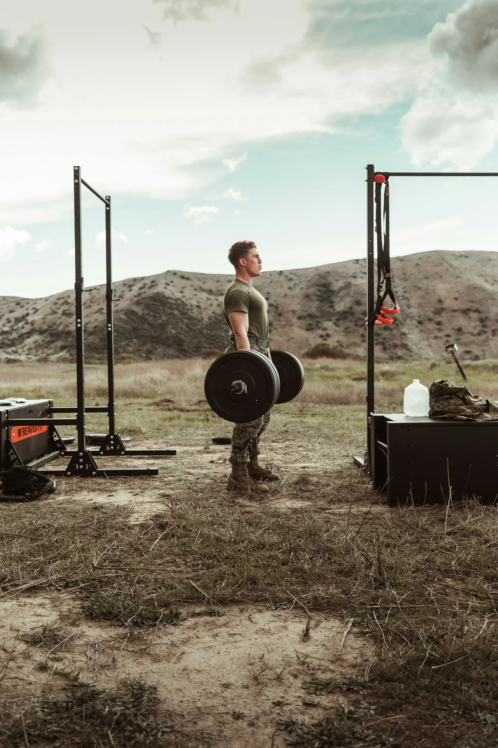 A man in military camouflage pants and a green t-shirt lifting weights outdoors in a field with hills in the background. Military tactical training in Grand Forks nd.