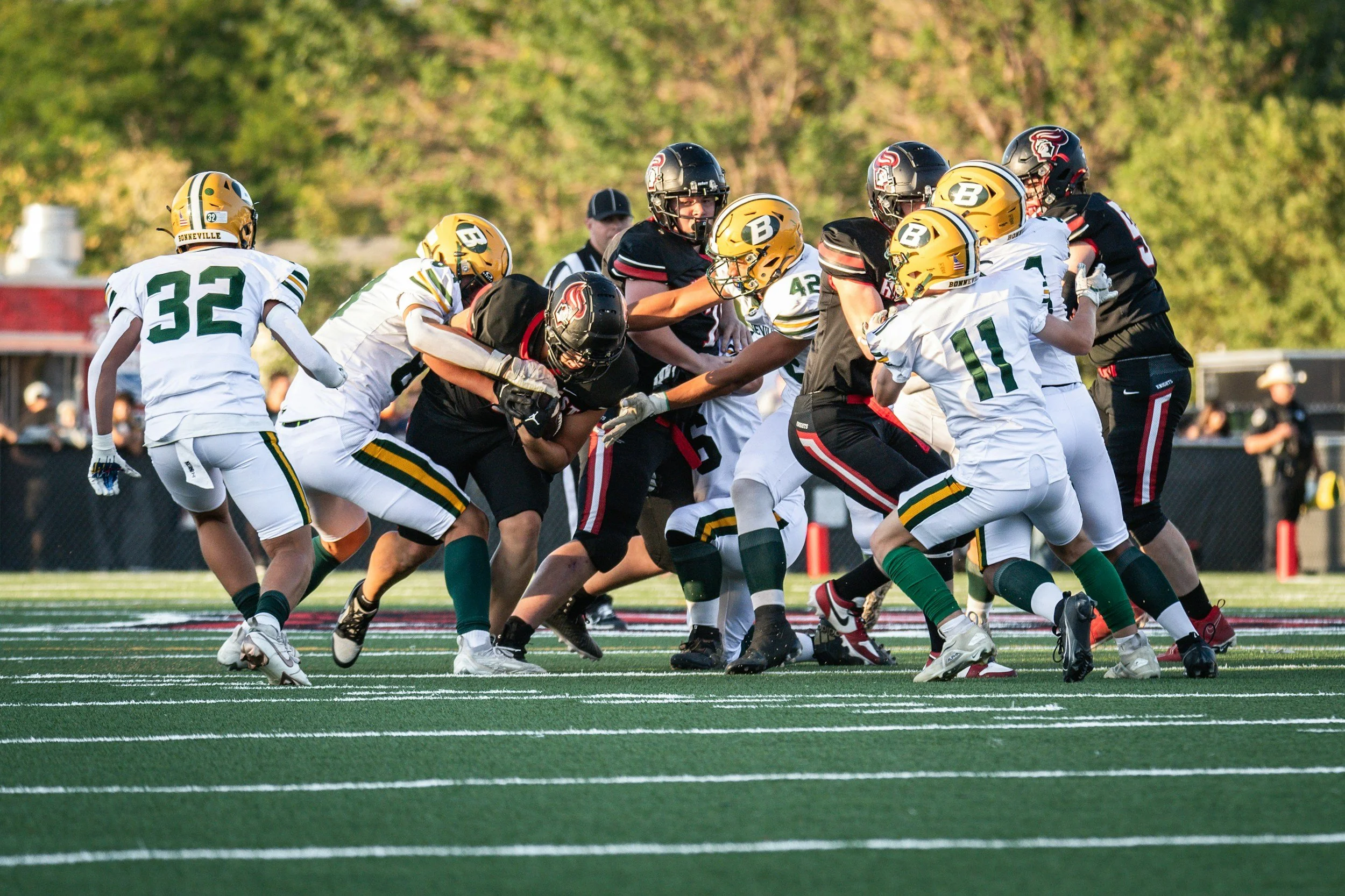 Young football players in black and white uniforms engage in a game on a green field with white yard lines, with trees and spectators in the background. Youth athlete training.