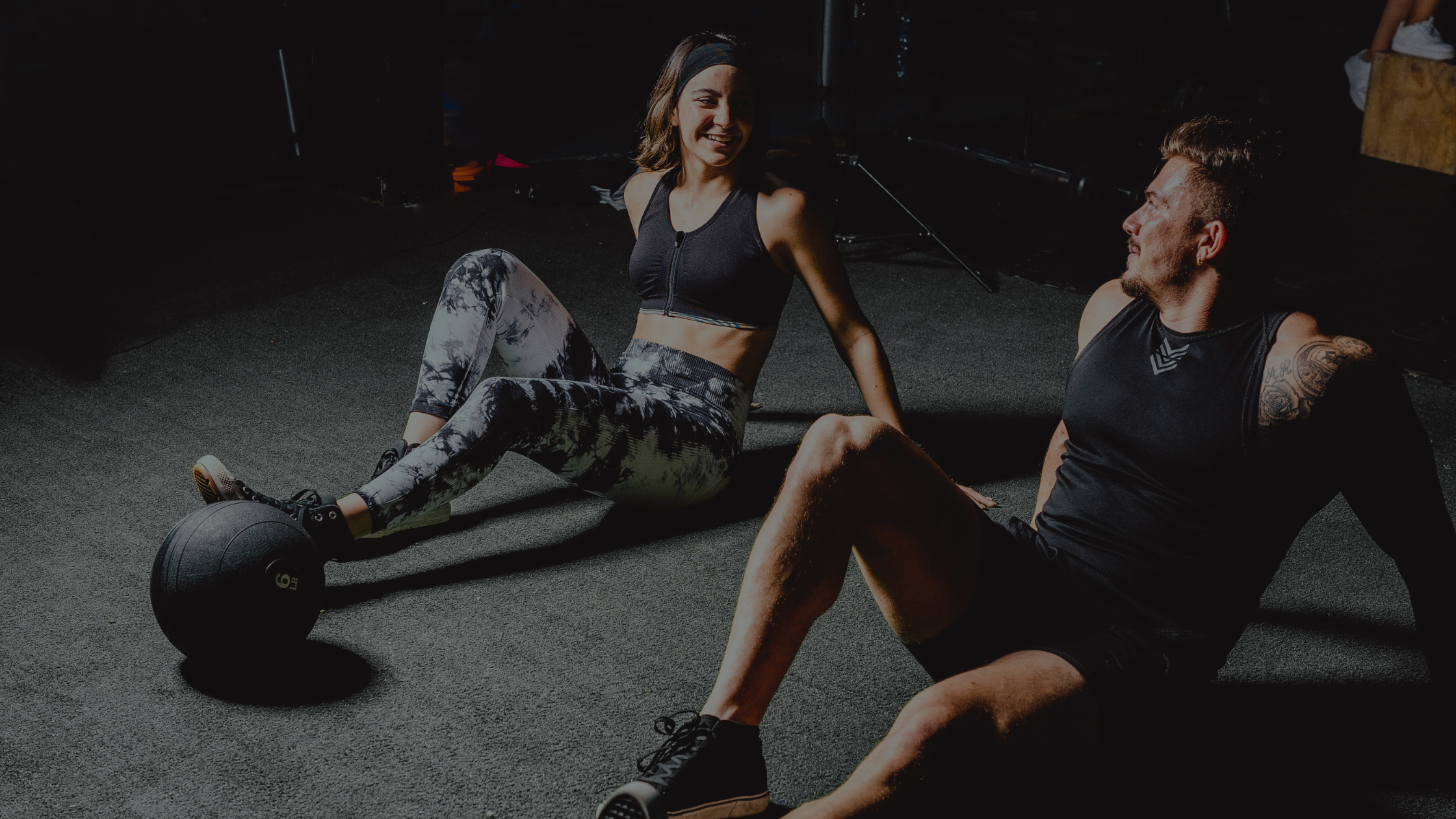 A woman and man sitting on a gym floor, smiling, after workout, with a kettlebell nearby.