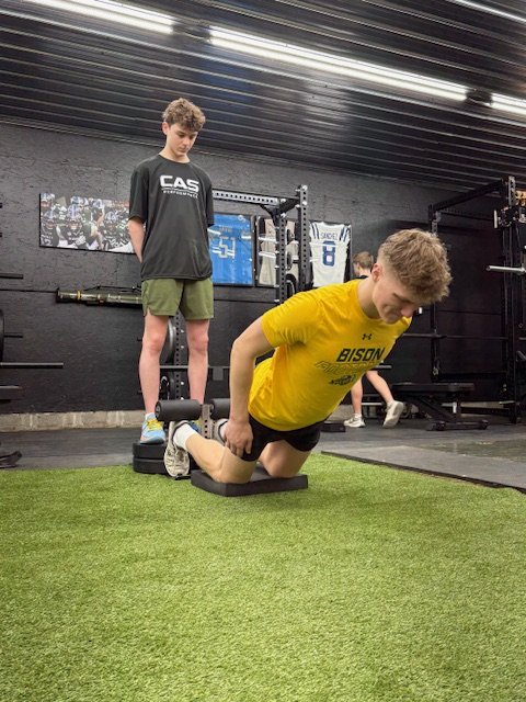 A young man in a yellow shirt is performing a stretch on a foam roller in a gym, with a person in a black shirt and shorts observing nearby.