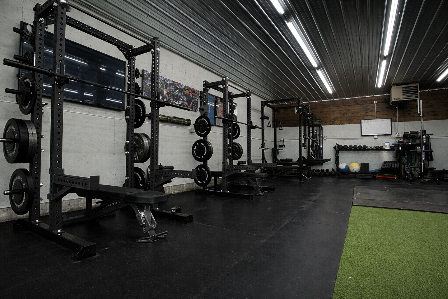 Interior of a gym with black workout equipment, including squat racks with weights, a bench, and various fitness accessories, with a green turf area on the right.