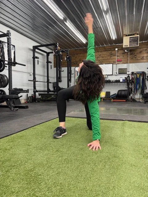 Woman performing a yoga pose on a green mat in a gym or fitness studio, with gym equipment and workout gear in the background.