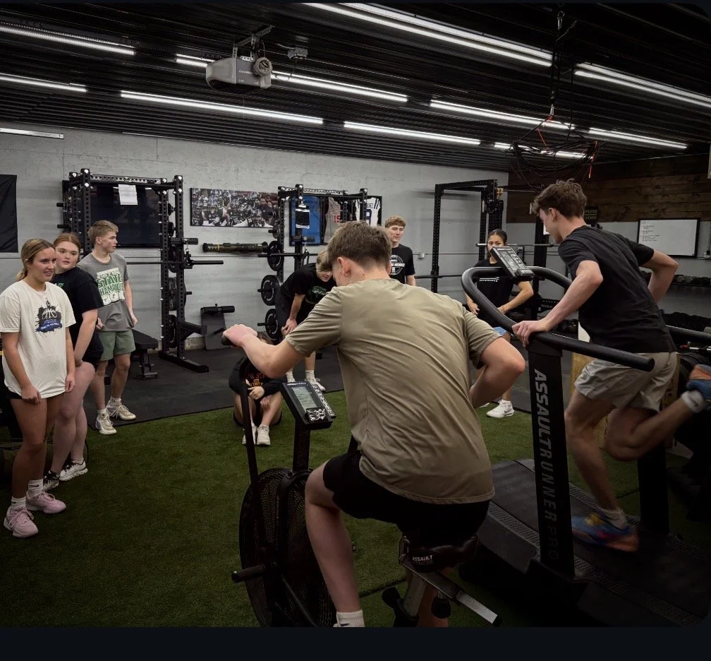 Group of young people at a gym with fitness equipment, two individuals on exercise bikes, others standing nearby watching. High school athlete training in grand forks.