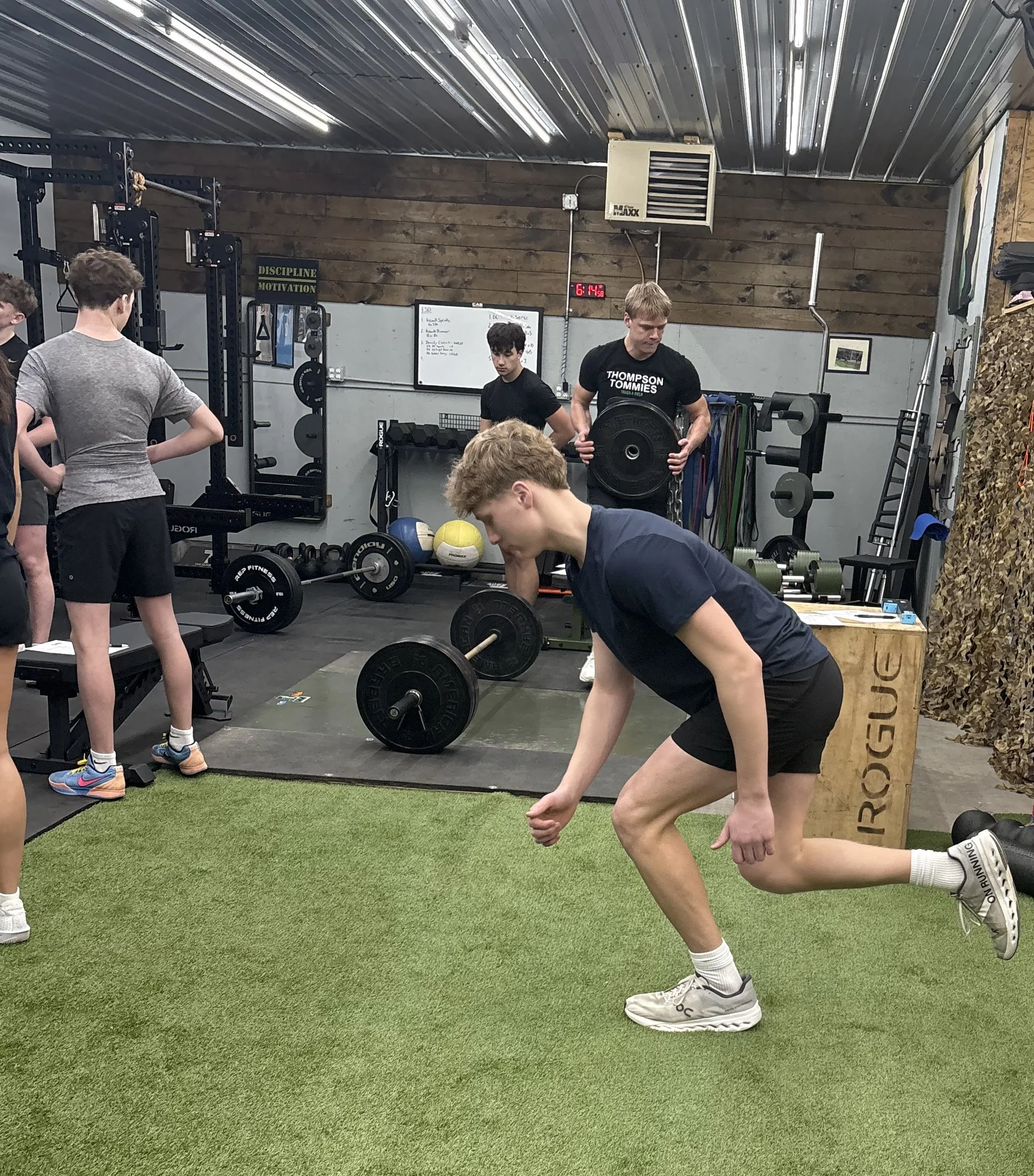 Young man performing a lunge exercise on a green turf floor in a gym, with weightlifting equipment and other people working out in the background.