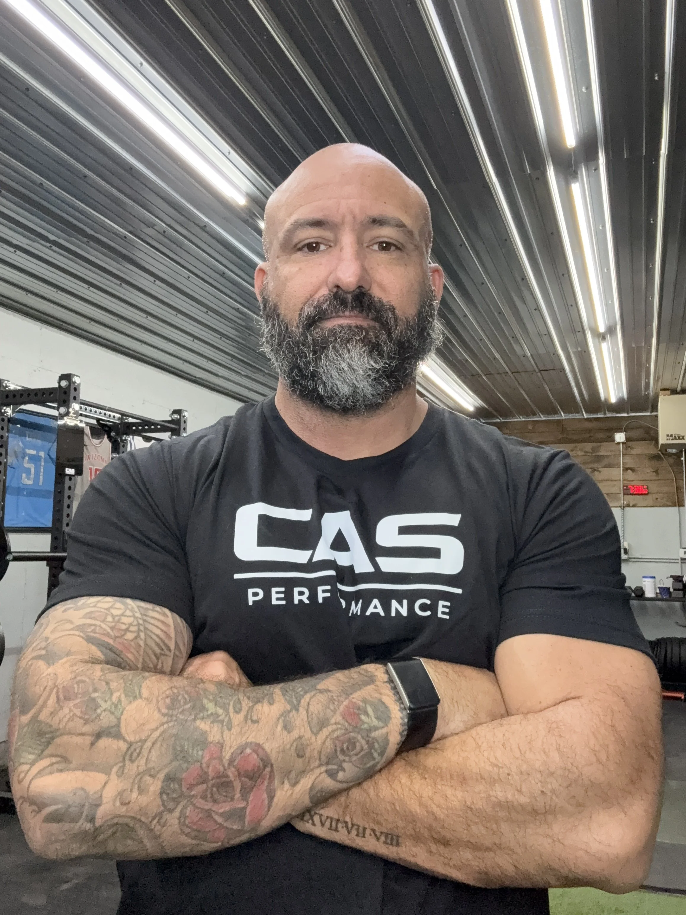 A personal trainer grand forks nd, crossing his arms, wearing a black CAS Performance t-shirt and a smartwatch, in a gym with metal ceiling and gym equipment in the background.