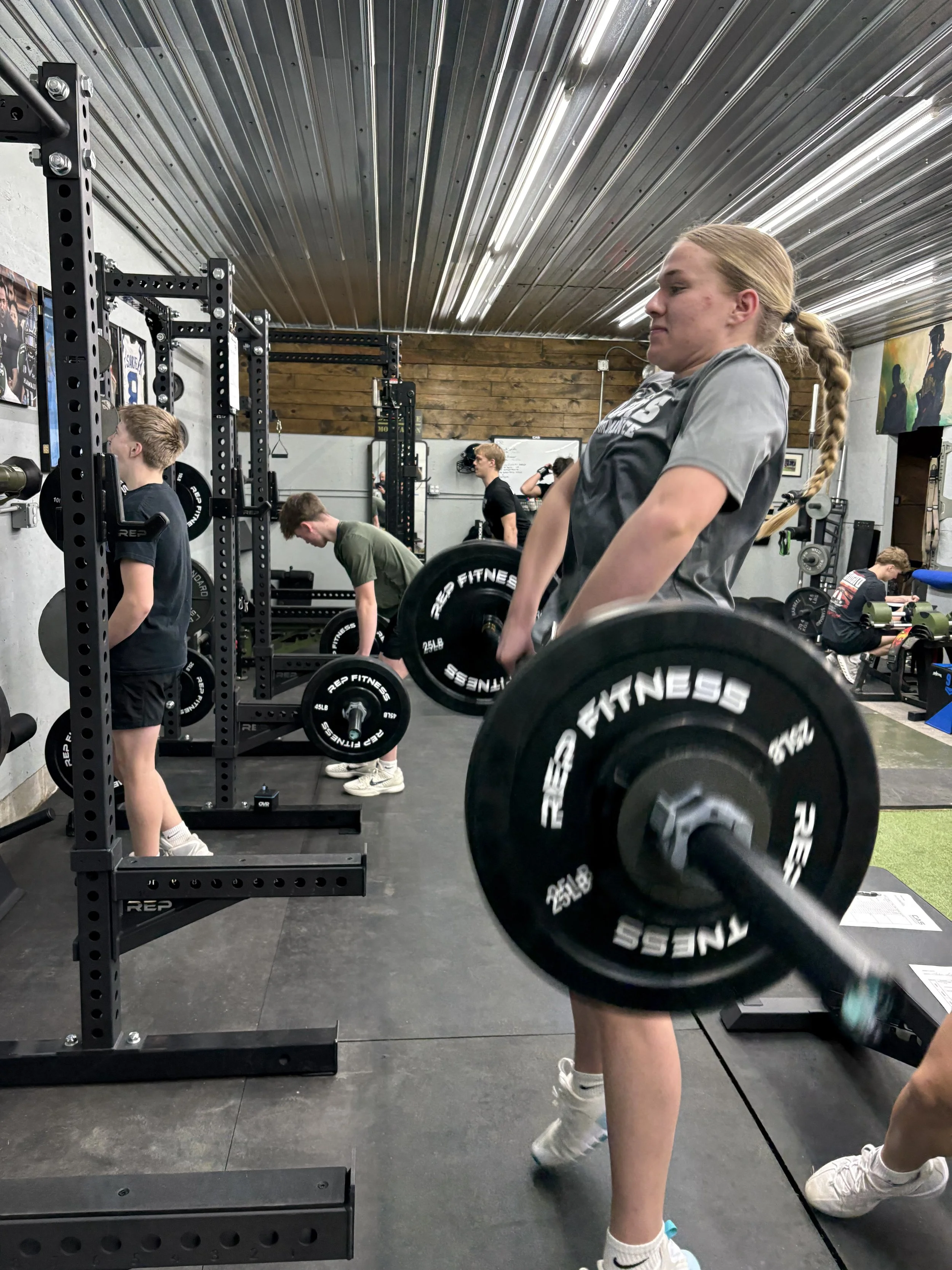 A young woman with a braid lifting a barbell at a gym, surrounded by other people working out.