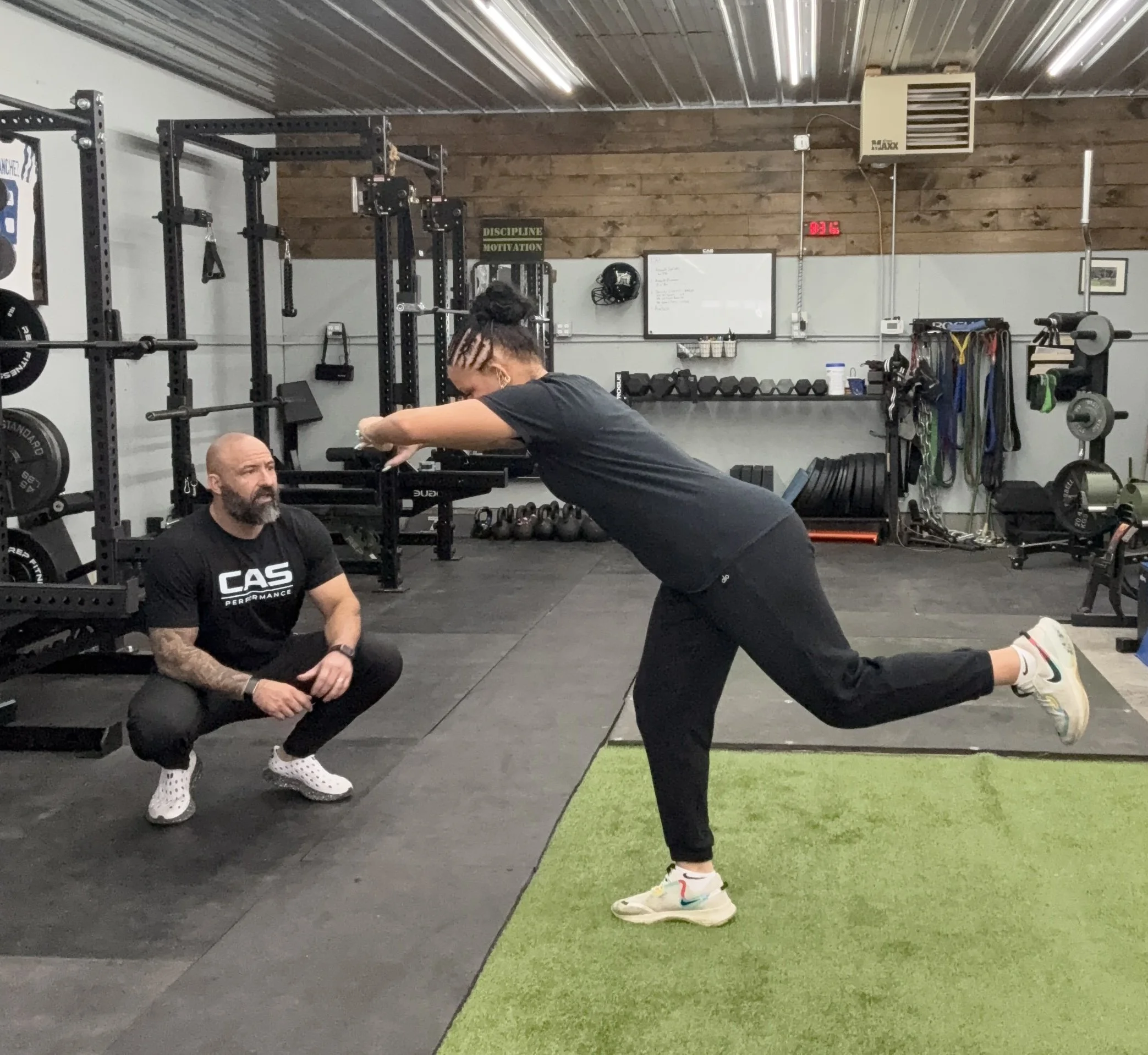 A woman performing a balance exercise with one leg raised and arms extended, supervised by a man crouching and observing, in a gym setting with workout equipment and weights.
