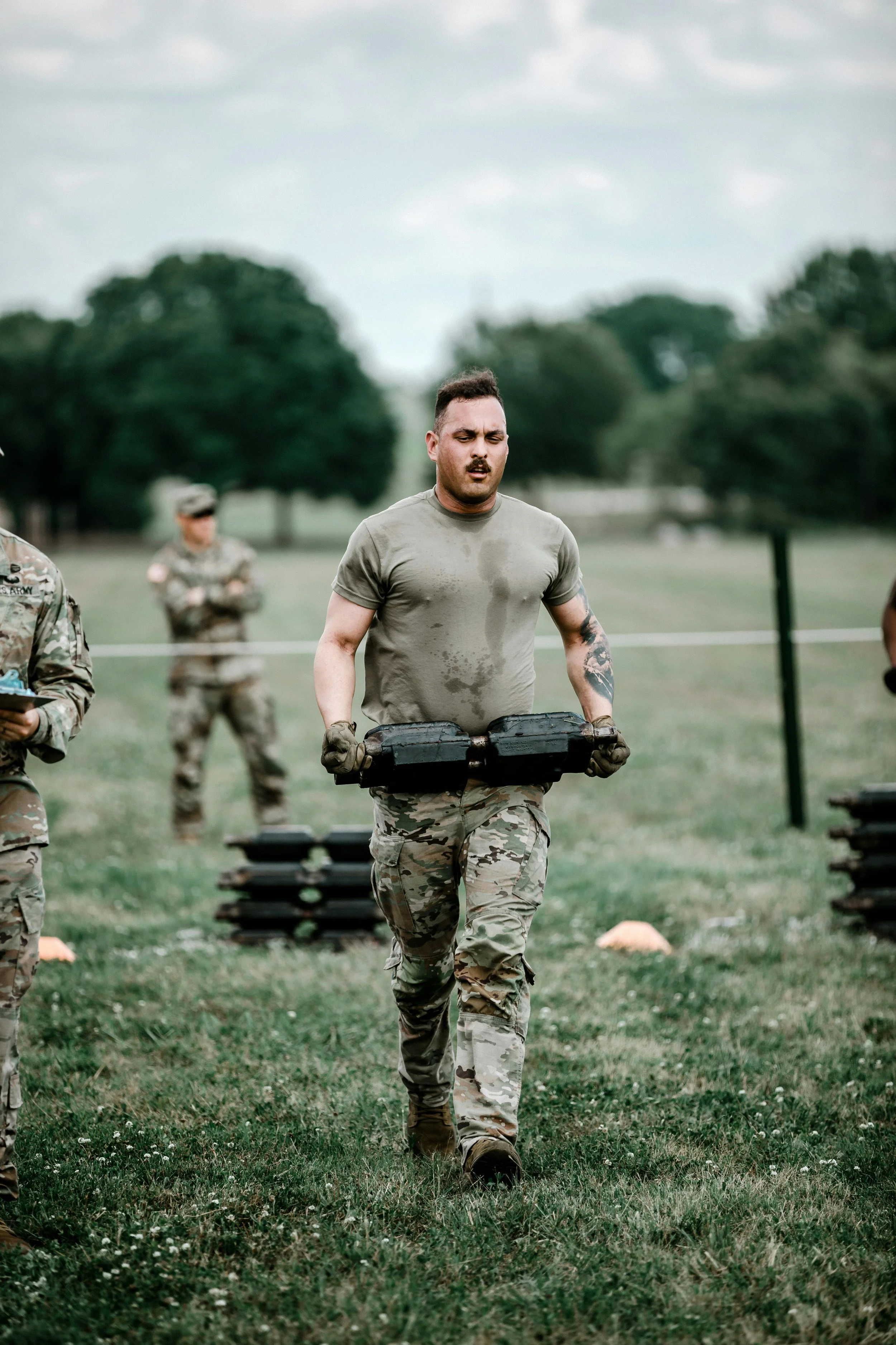 A soldier in camouflage uniform carrying a black box during a military training exercise on a grassy field, with other soldiers in the background.