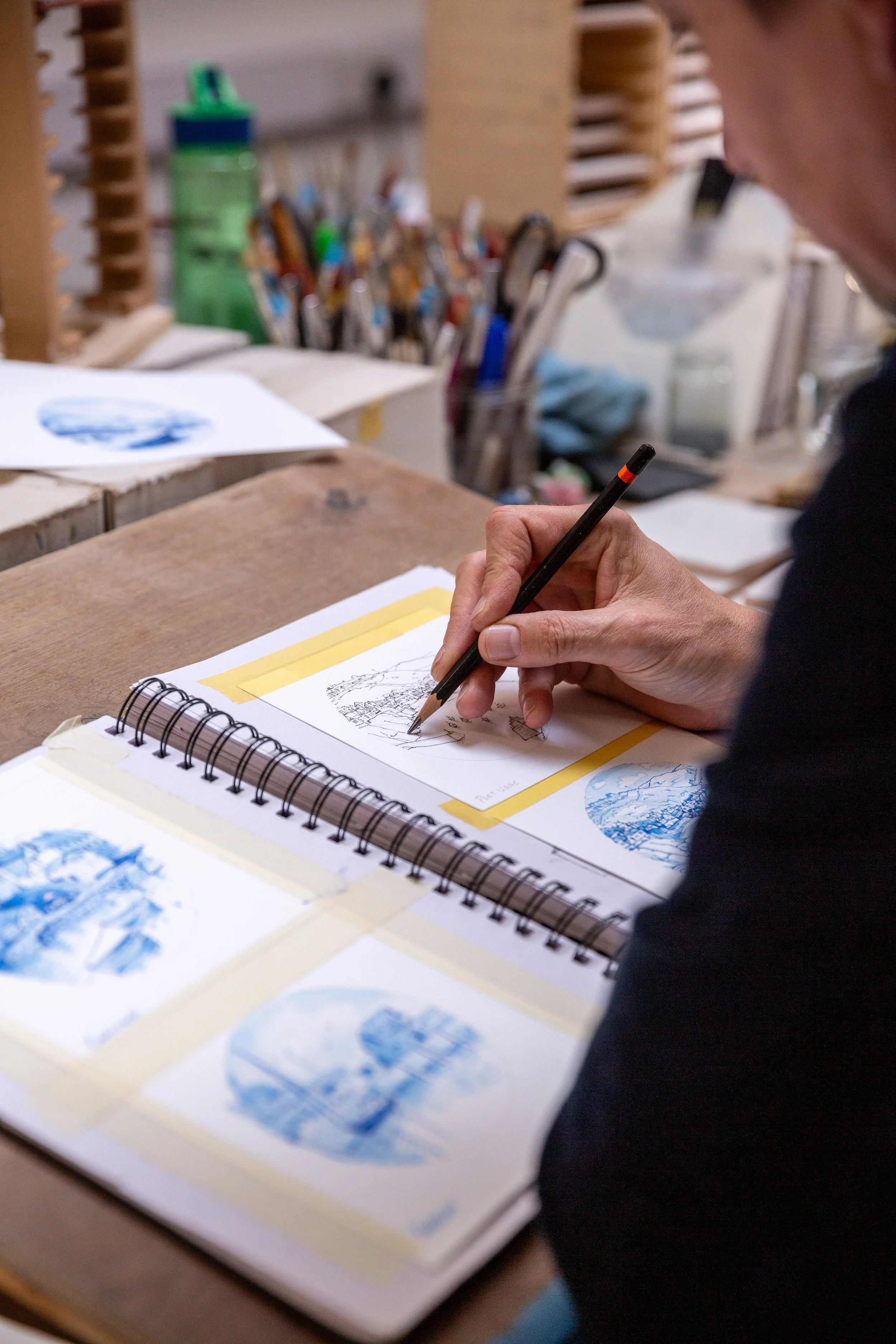 A person sketches in a notebook surrounded by printed blue landscape images, sitting at a wooden desk in an art studio with various art supplies and tools in the background.