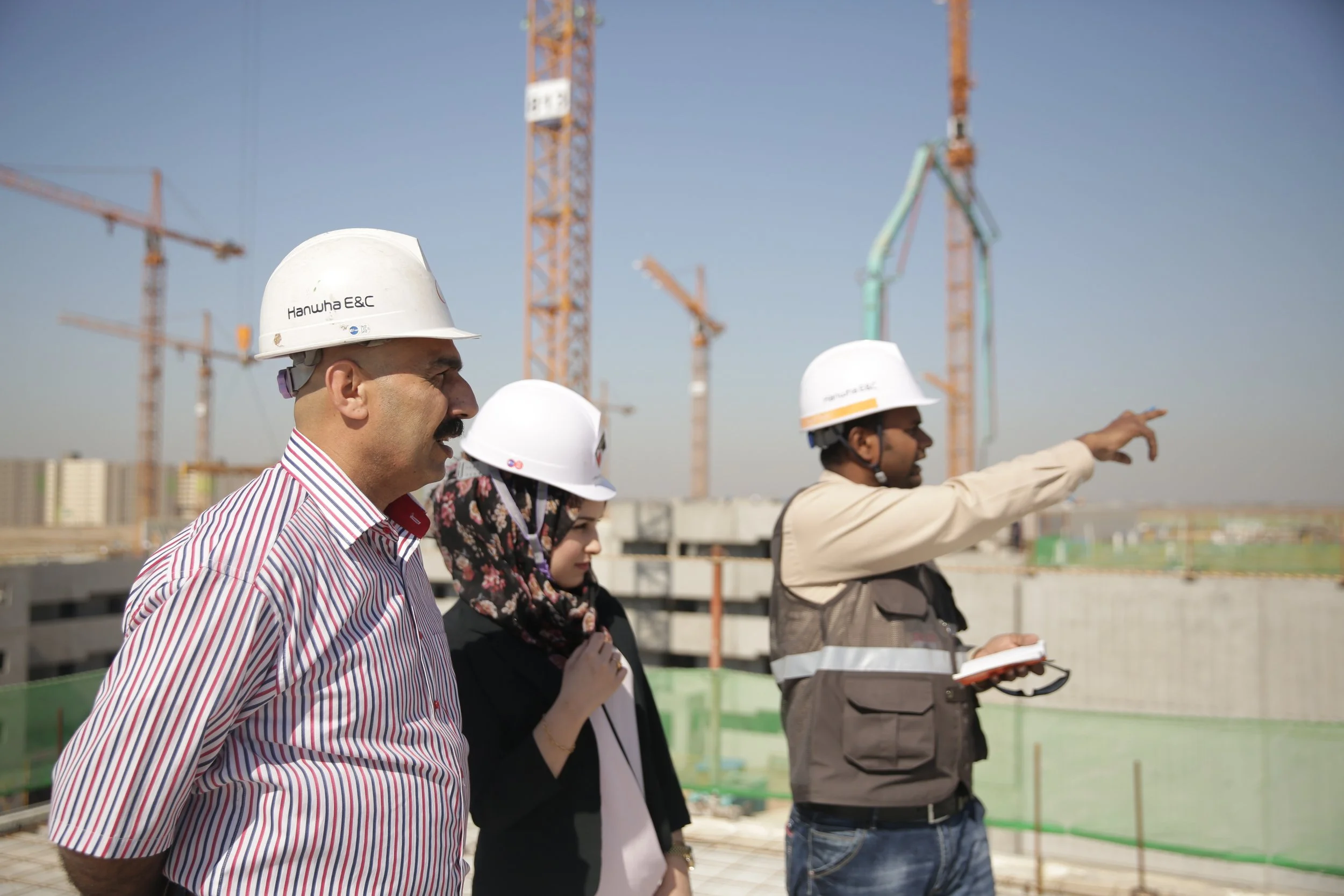 Three construction professionals on a building site, wearing hard hats, with cranes and a construction structure in the background. One person is pointing towards the structure while holding a notebook.