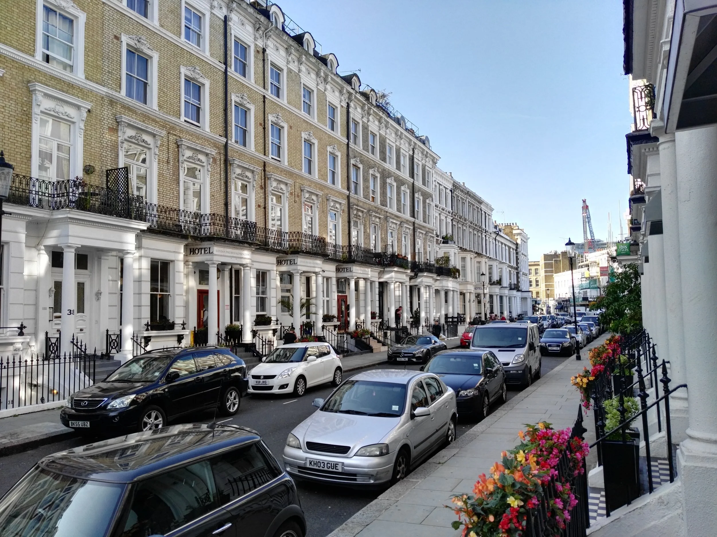 Streetside view of a row of historic white and beige townhouses with black wrought-iron balconies, parked cars along the curb, and colorful flowers in window boxes and on small front steps under a blue sky.