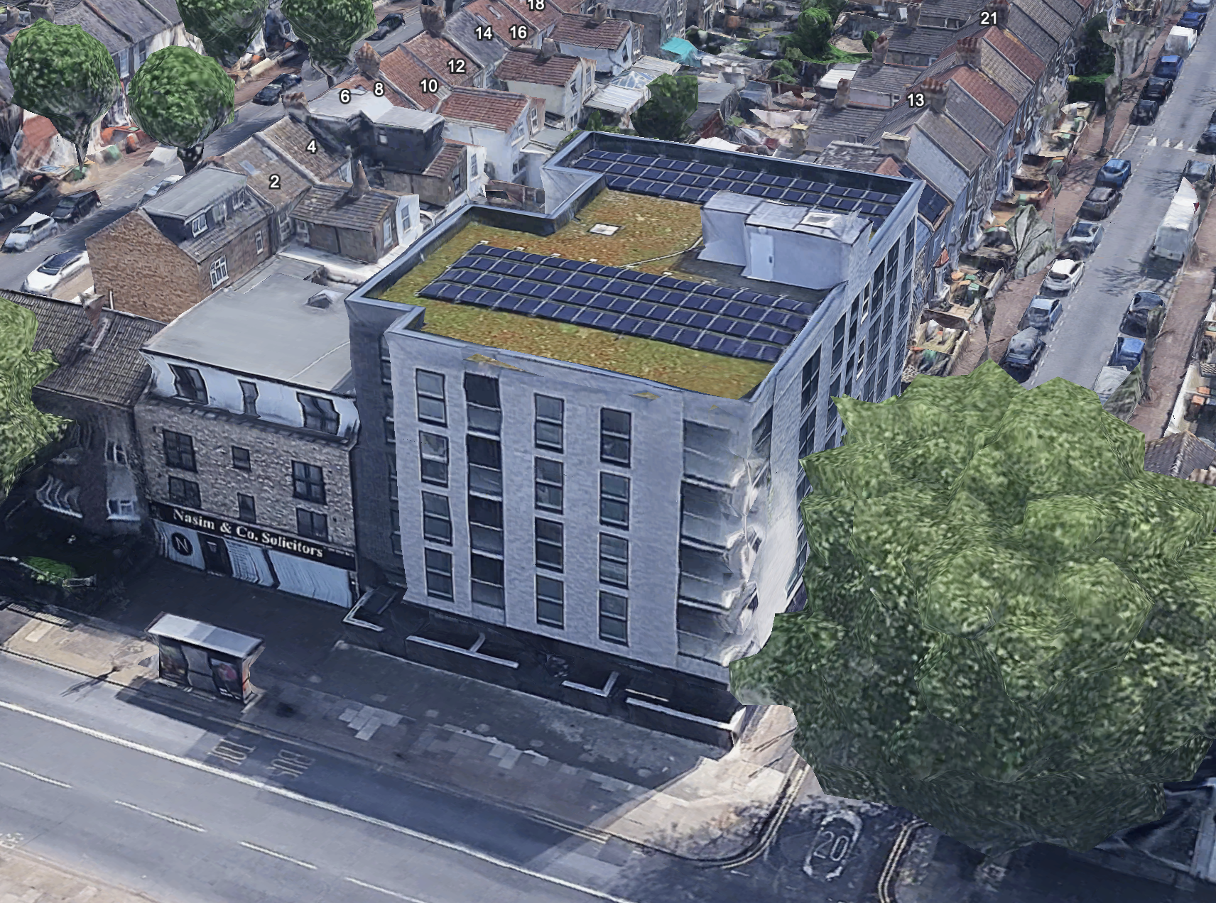 Aerial view of a modern multi-story building with solar panels on its roof, located in a neighborhood with small, older houses and a parking lot, next to a tree-lined street.