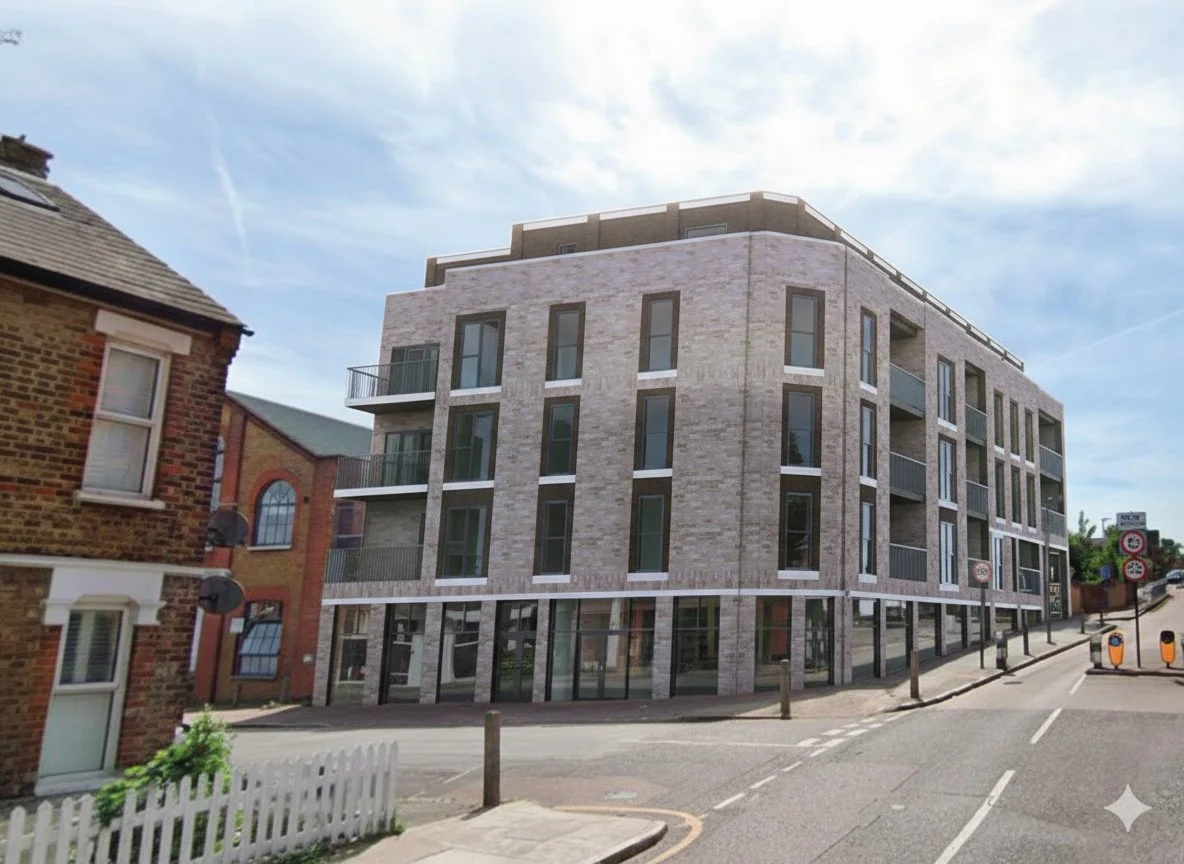 A modern, multi-story residential building with brick and glass exterior, situated at a street corner with a sidewalk and traffic barriers.