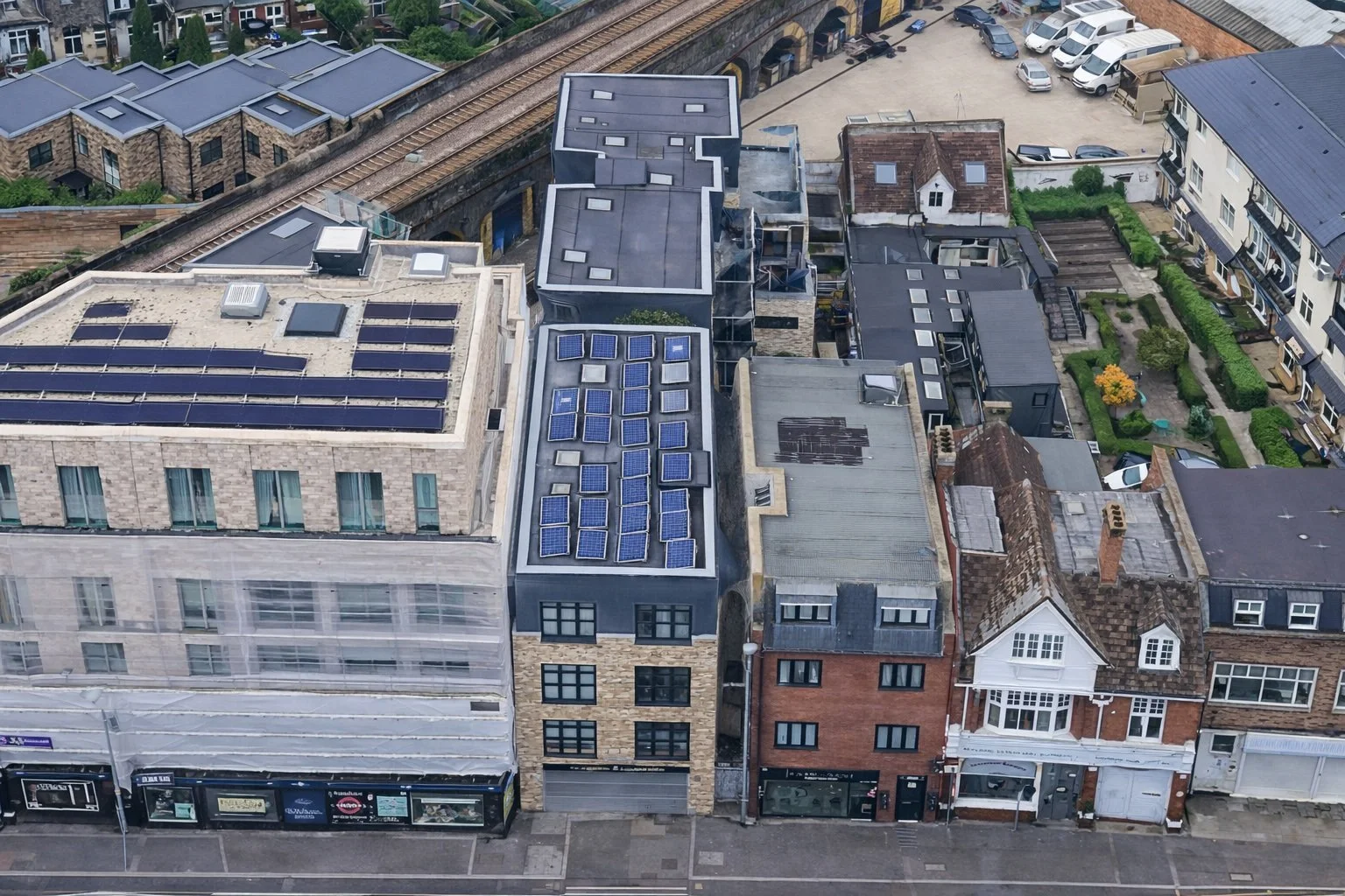 Aerial view of urban buildings with solar panels on rooftops, adjacent to a railway track, with a street at the bottom lined with shops.