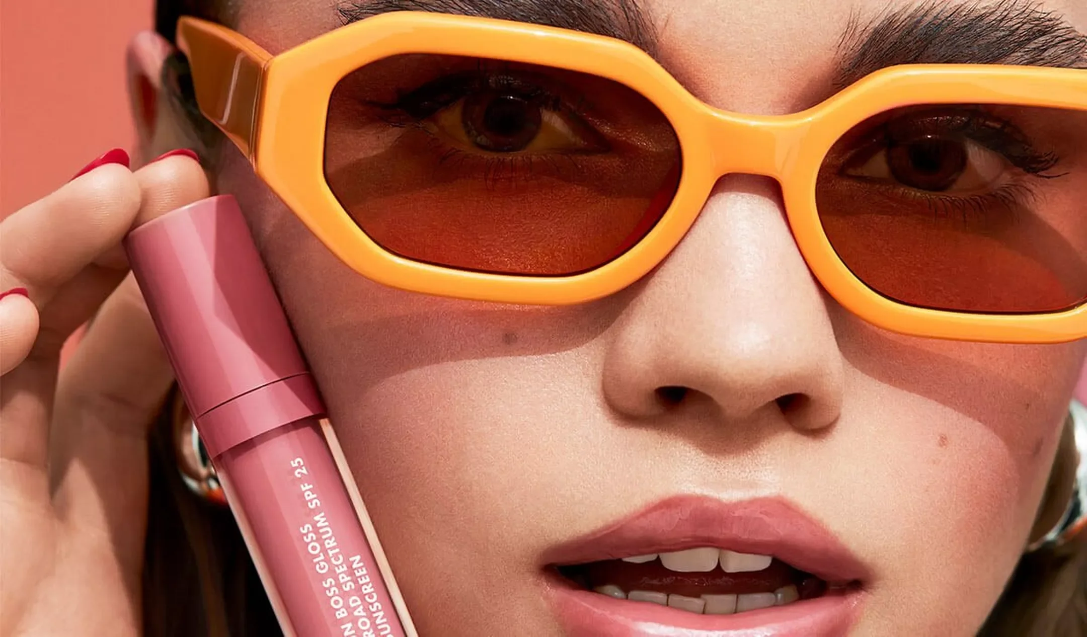 Close-up of a woman with fair skin wearing orange sunglasses with red-tinted lenses, holding a pink sunscreen tube near her cheek, with a pink background.