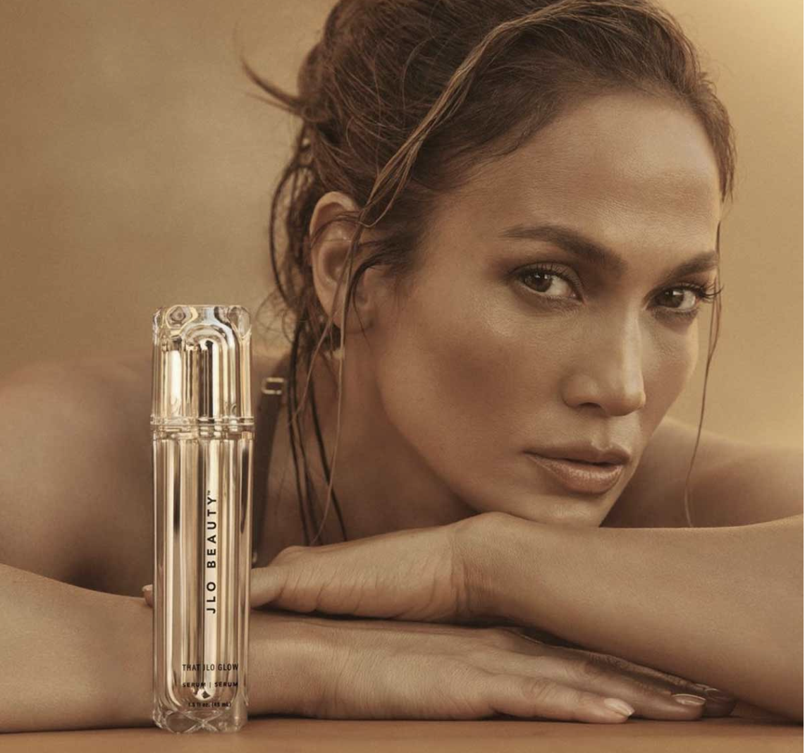 Jennifer Lopez with her head on her arms, featuring a skincare product labeled 'JLO BEAUTY' positioned in front of her. The background and overall tone are warm and soft.