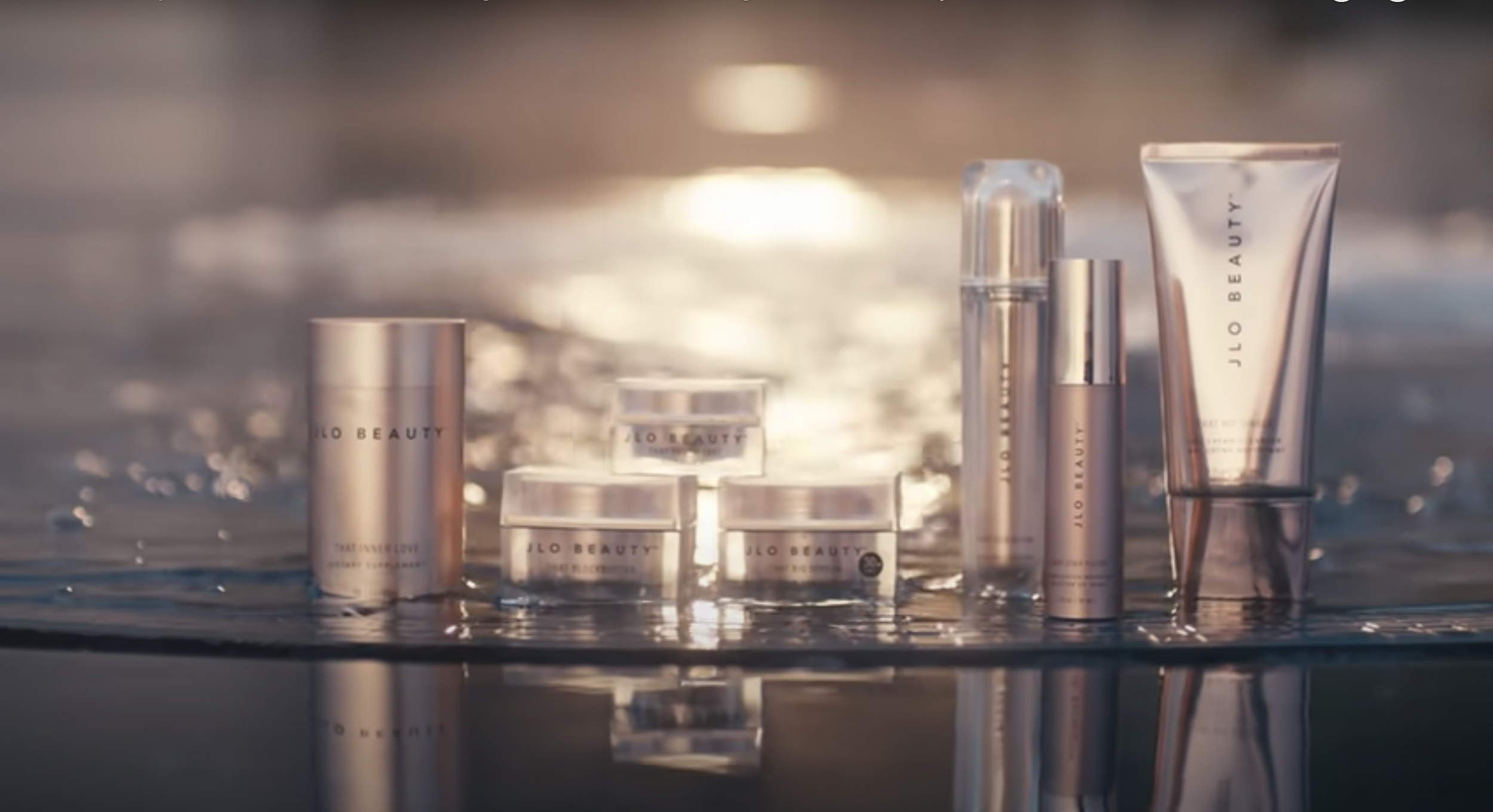 A collection of skincare products from 'JLo Beauty' placed on water with sunlight reflecting in the background.