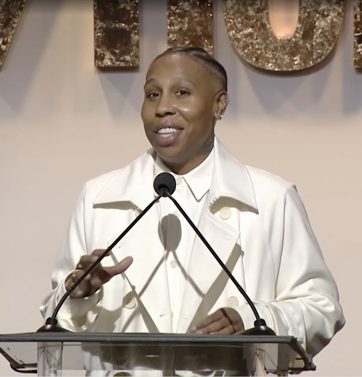 Lena Waithe speaking at a podium for Yahoo Makers Honors Awards night with a microphone, wearing a white jacket, with a decorative wall hanging in the background.