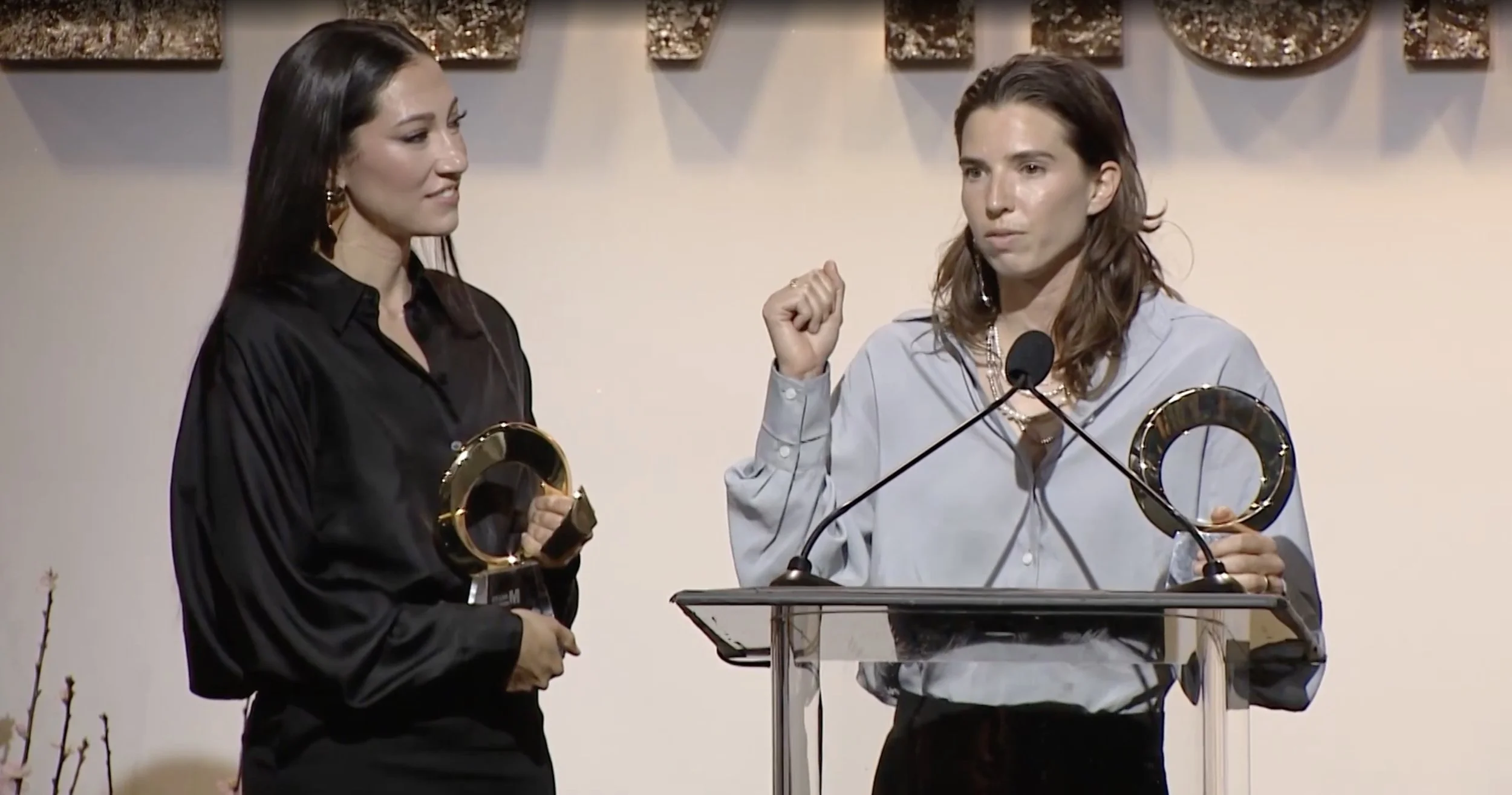 Christen Press on the left and Tobin Heath on the right standing at a podium for Yahoo's Makers Honor awards night, one speaking and the other holding an award for re-inc. Tobin Heatht appears to be making a fist gesture.