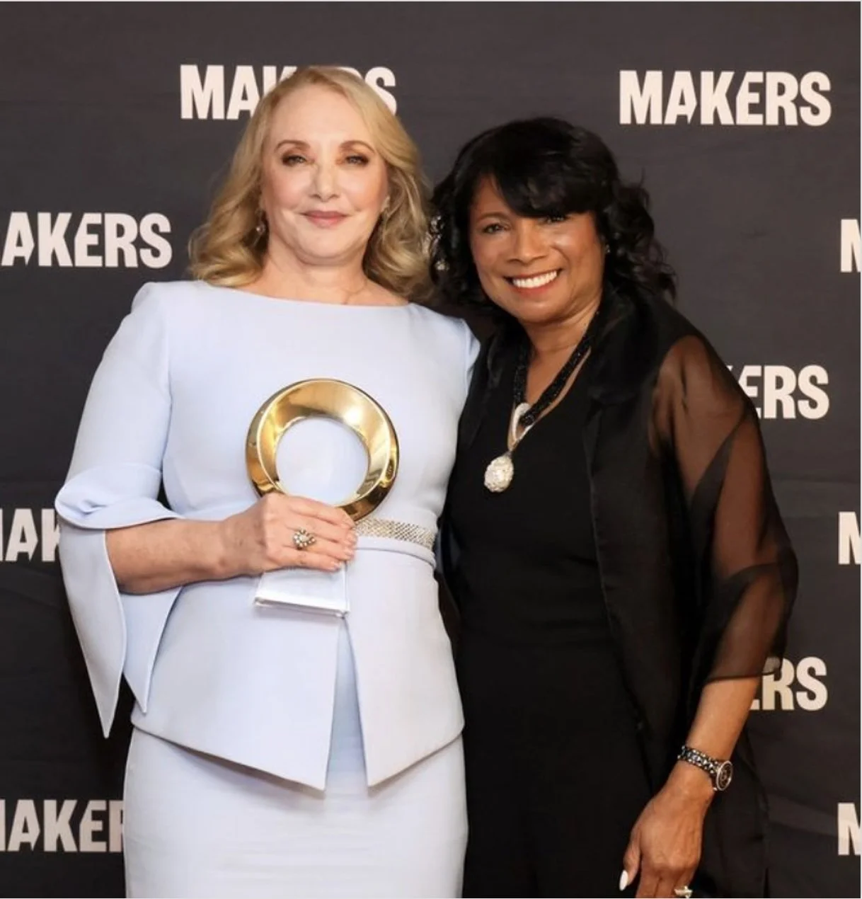 J. Smith Cameron standing close together with a black woman, smiling, at the Yahoo Makers Honors awards event with a backdrop that says 'Makers.' J. Smith Cameron is holding a round, gold trophy or award.
