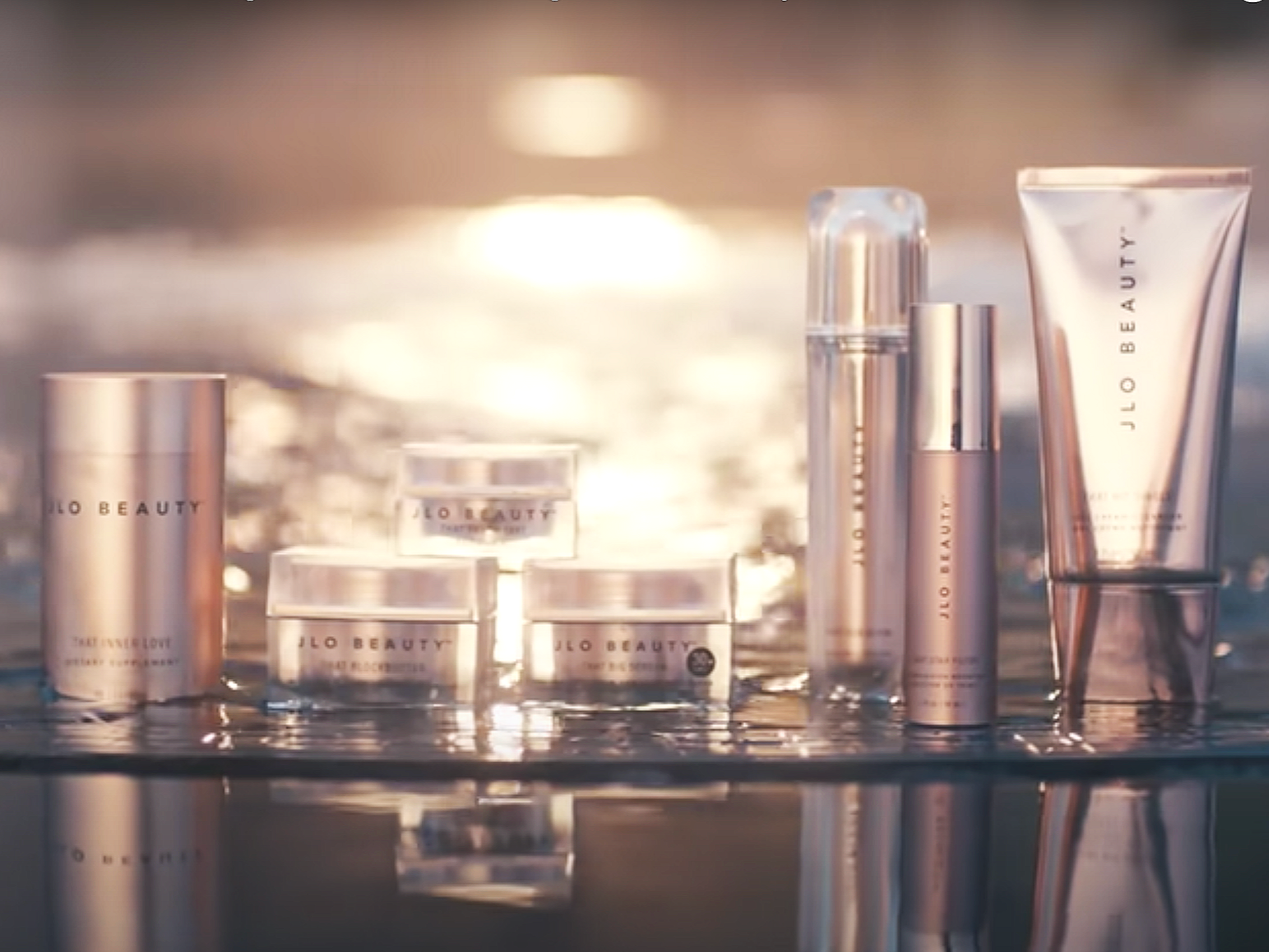 A collection of skincare products from 'JLo Beauty' placed on water with sunlight reflecting in the background.