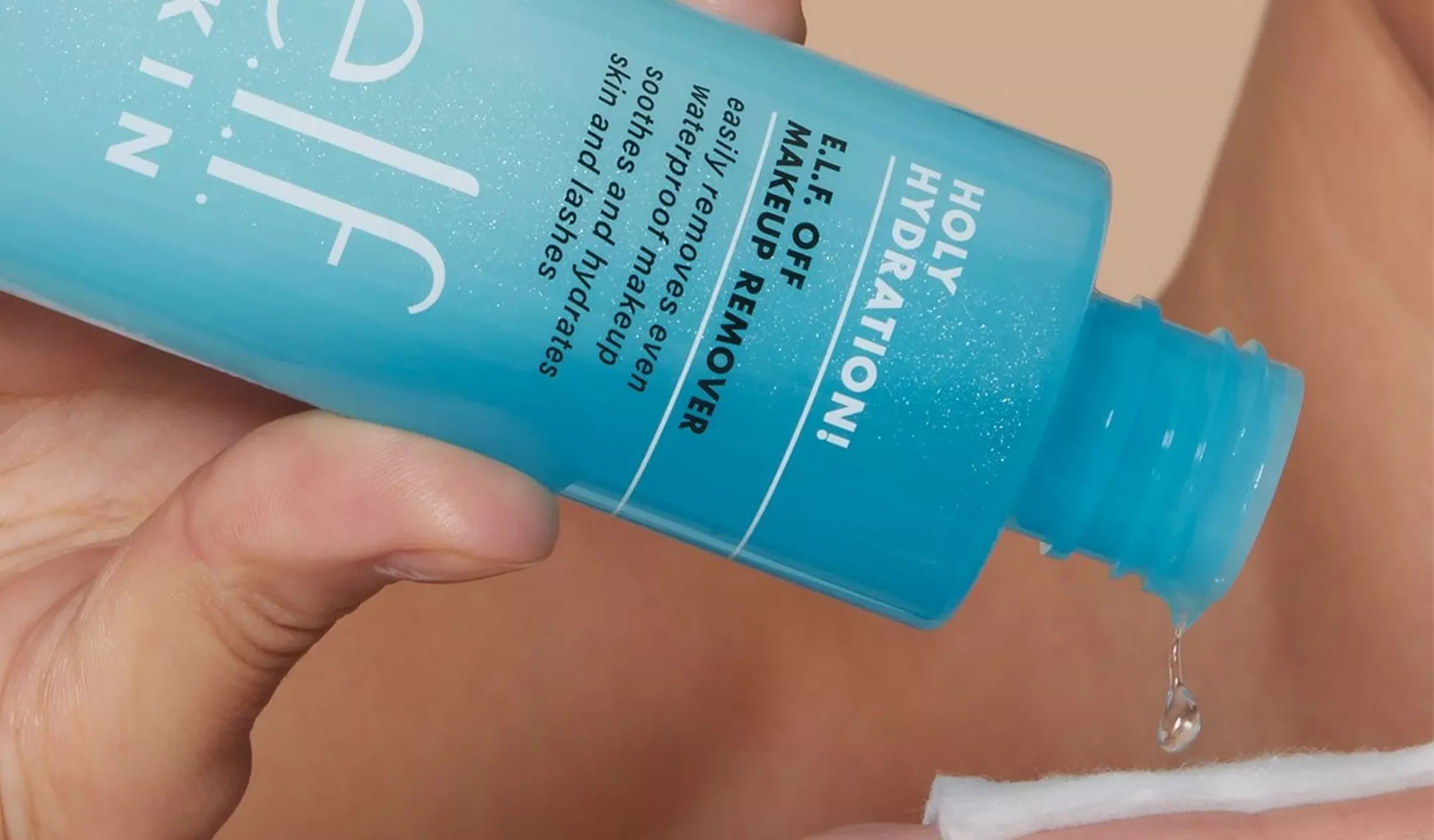 A hand holding a blue bottle of Holy Hydration! ELLE Off Makeup Remover, with a drop of liquid falling from the opening onto a white cotton pad.