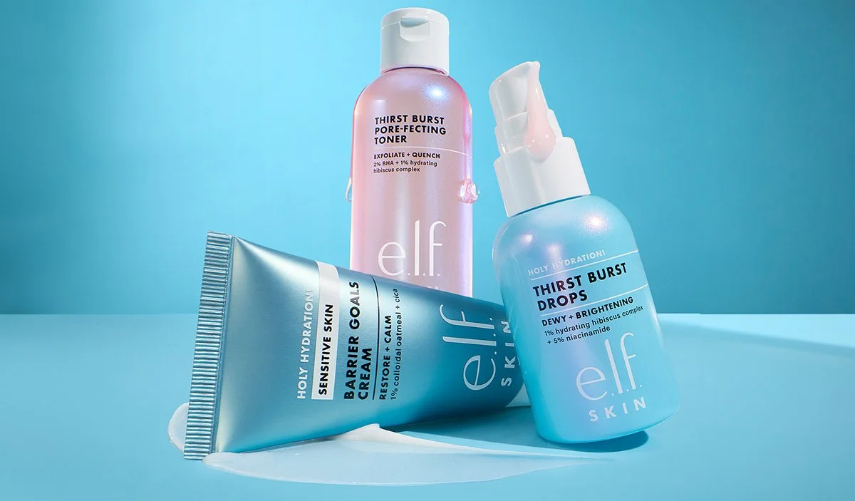 Three bottles of e.l.f. skincare products on a light blue surface with a blue background. One is a pink bottle of pore-fecting toner, another a blue bottle of drops, and the third a tube of sensitive skin barrier cream.