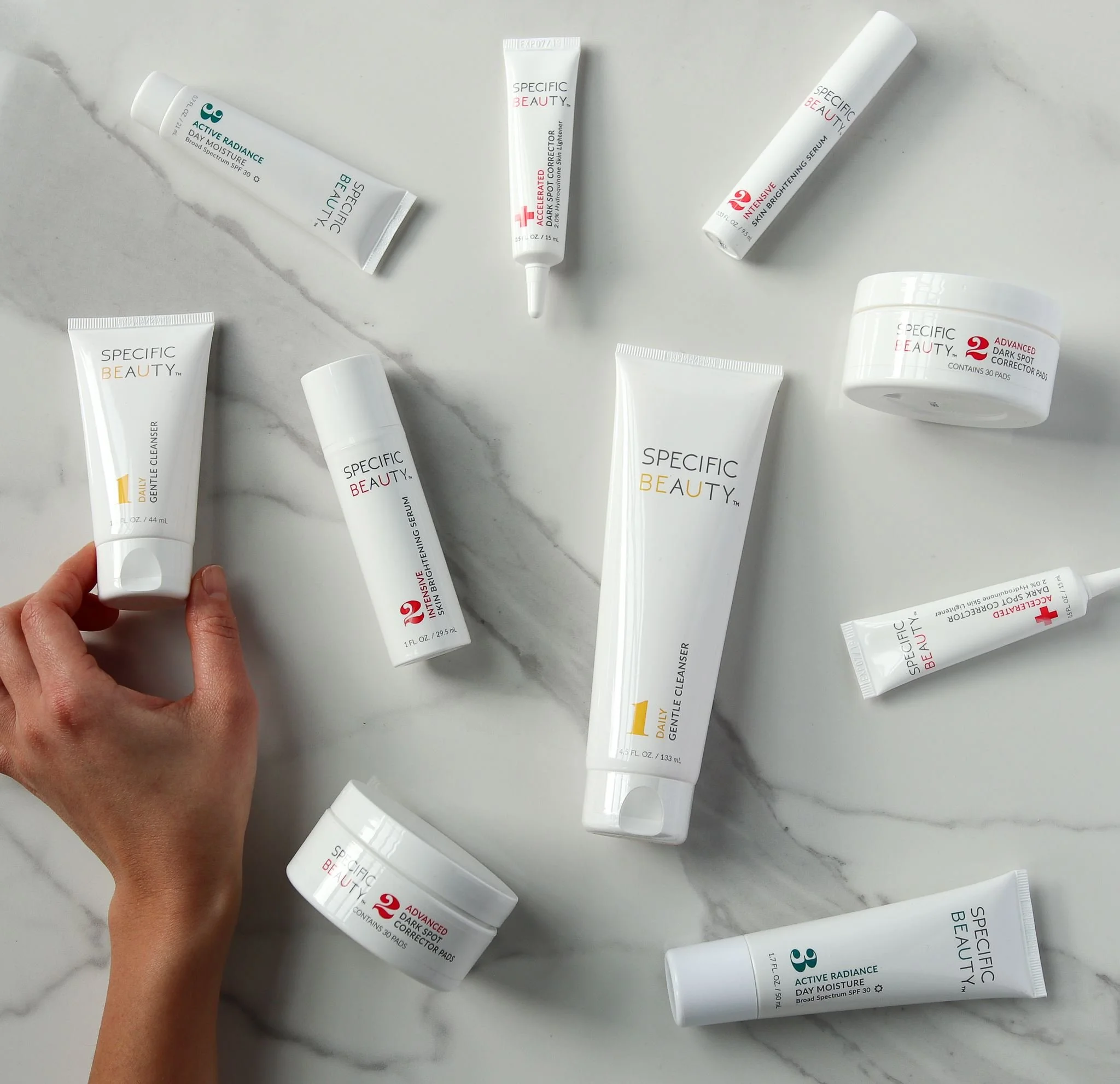 Collection of skincare products from Specific Beauty, including cleansers, serums, sunscreens, and treatment creams, arranged on a marble surface with one hand holding a tube of daily gentle cleanser.