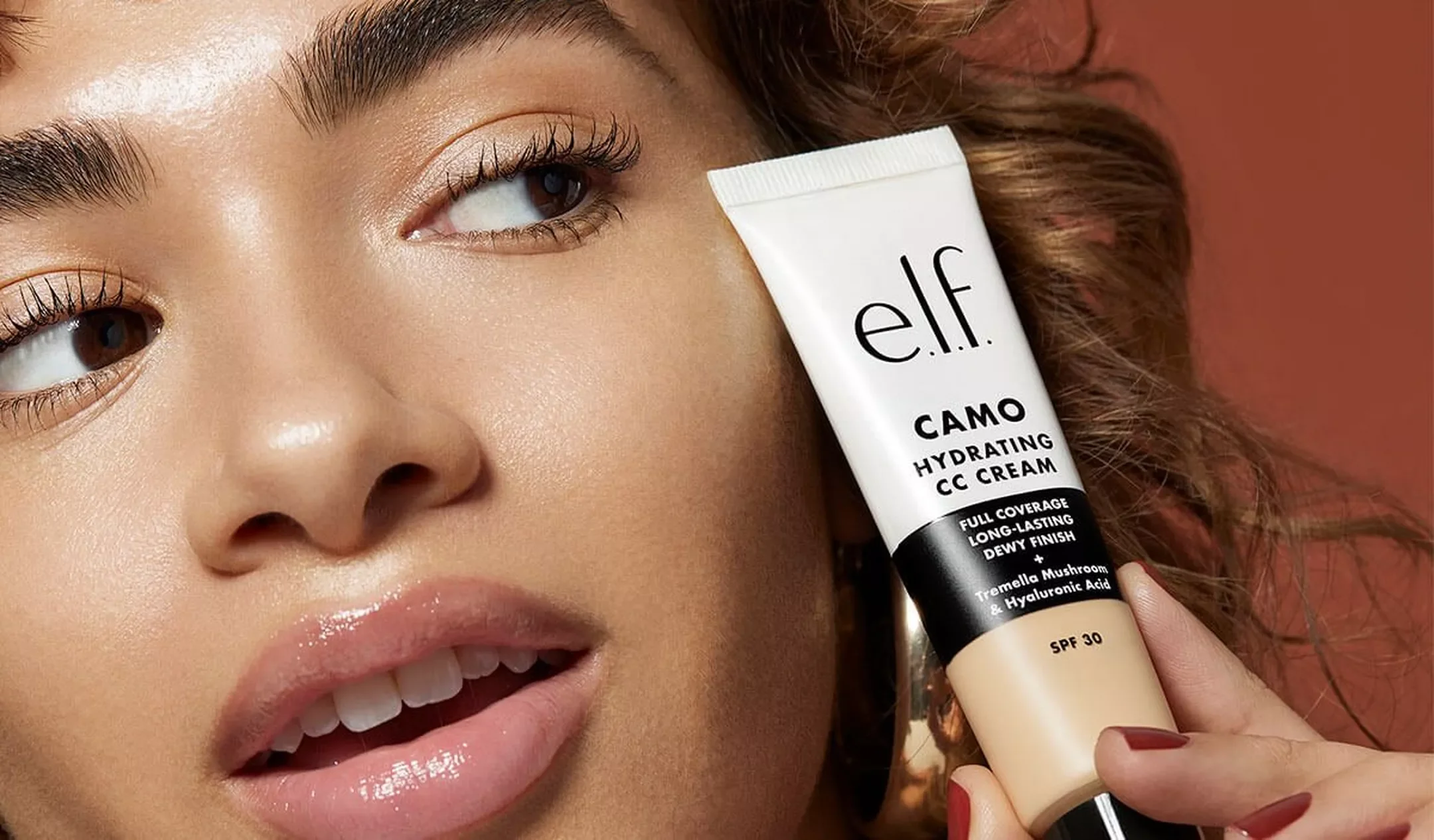 A woman with smooth skin holding a tube of e.l.f. CAMO Hydrating CC Cream near her face. The focus is on her face and the skincare product in a close-up shot.