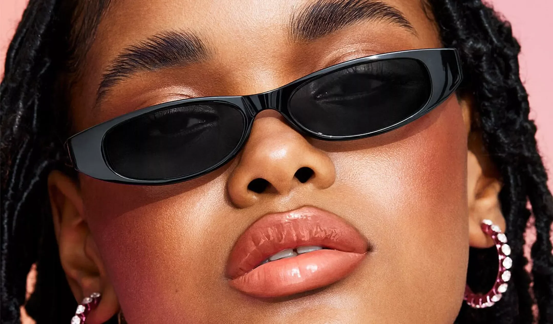 Close-up of a woman wearing black sunglasses with a pink background.