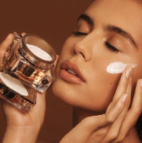 A woman applying cream to her face with her eyes closed, holding a jar of skincare product.