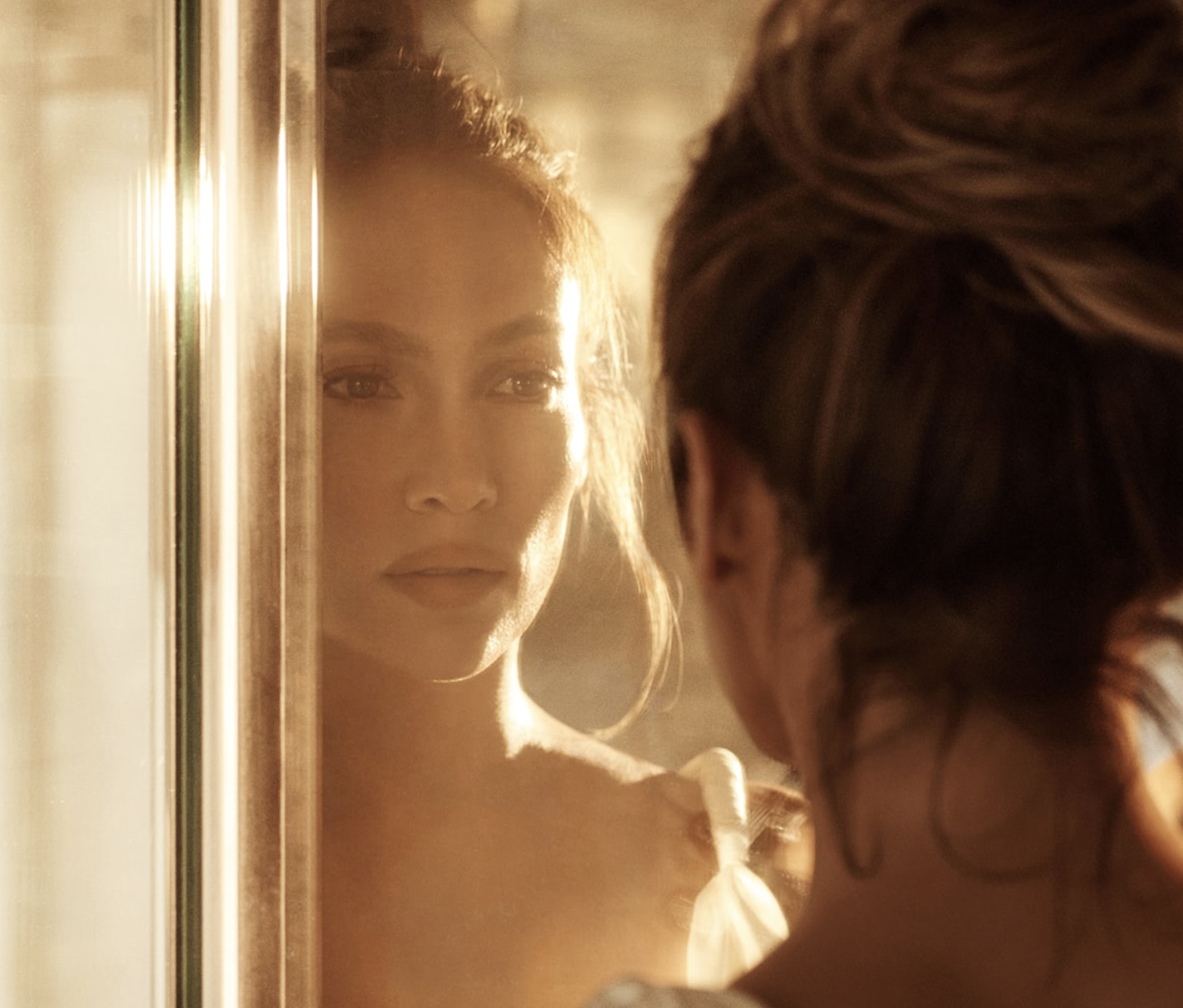 Jennifer Lopez looking at herself in a mirror with warm lighting, with her reflection in focus and her back to the camera shot for 'JLO Beauty'.