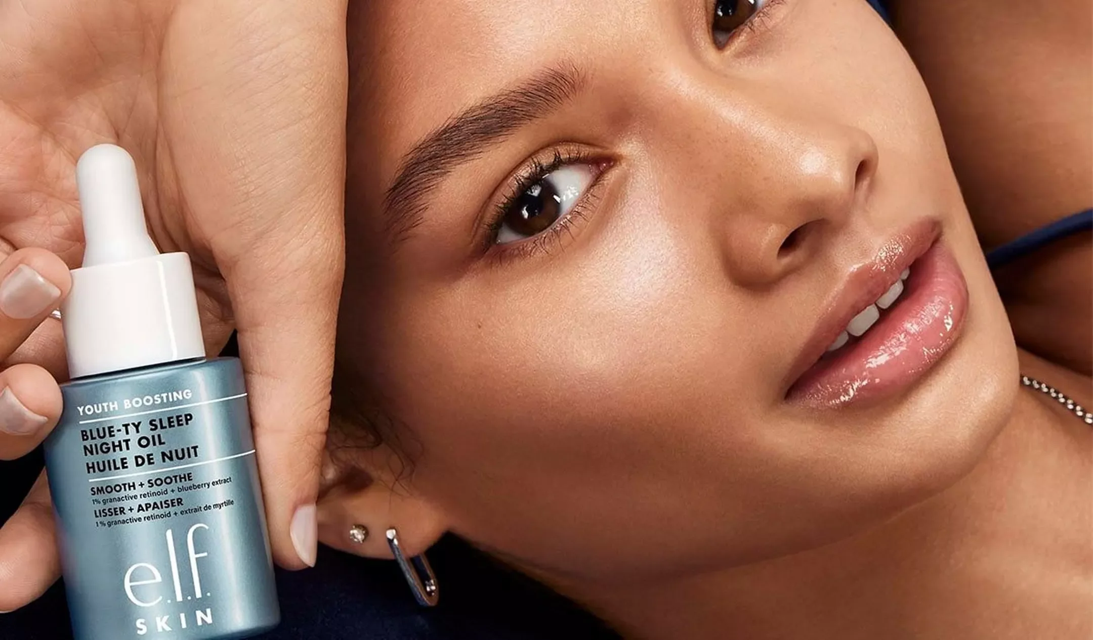 Close-up of a woman's face with smooth skin, lips slightly parted, lying down with her head resting on her hand, holding a bottle of elf sleep oil near her face.